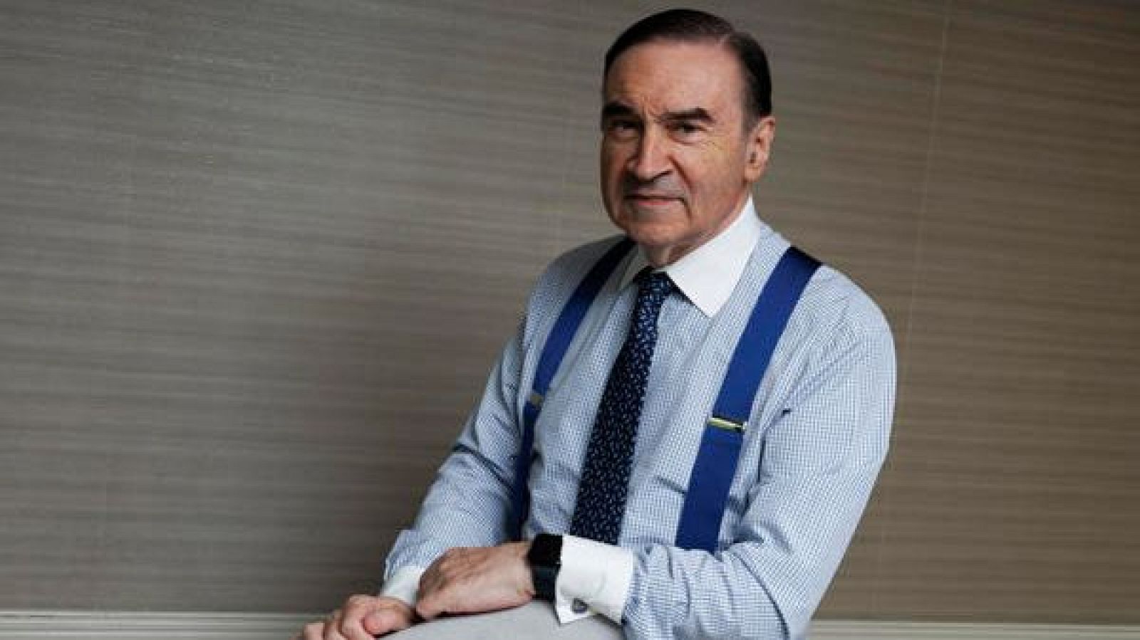 Pedro J. Ramírez, con camisa a cuadros, corbata oscura y tirantes azules, posa frente a una pared lisa. Lleva un reloj en la muñeca izquierda.