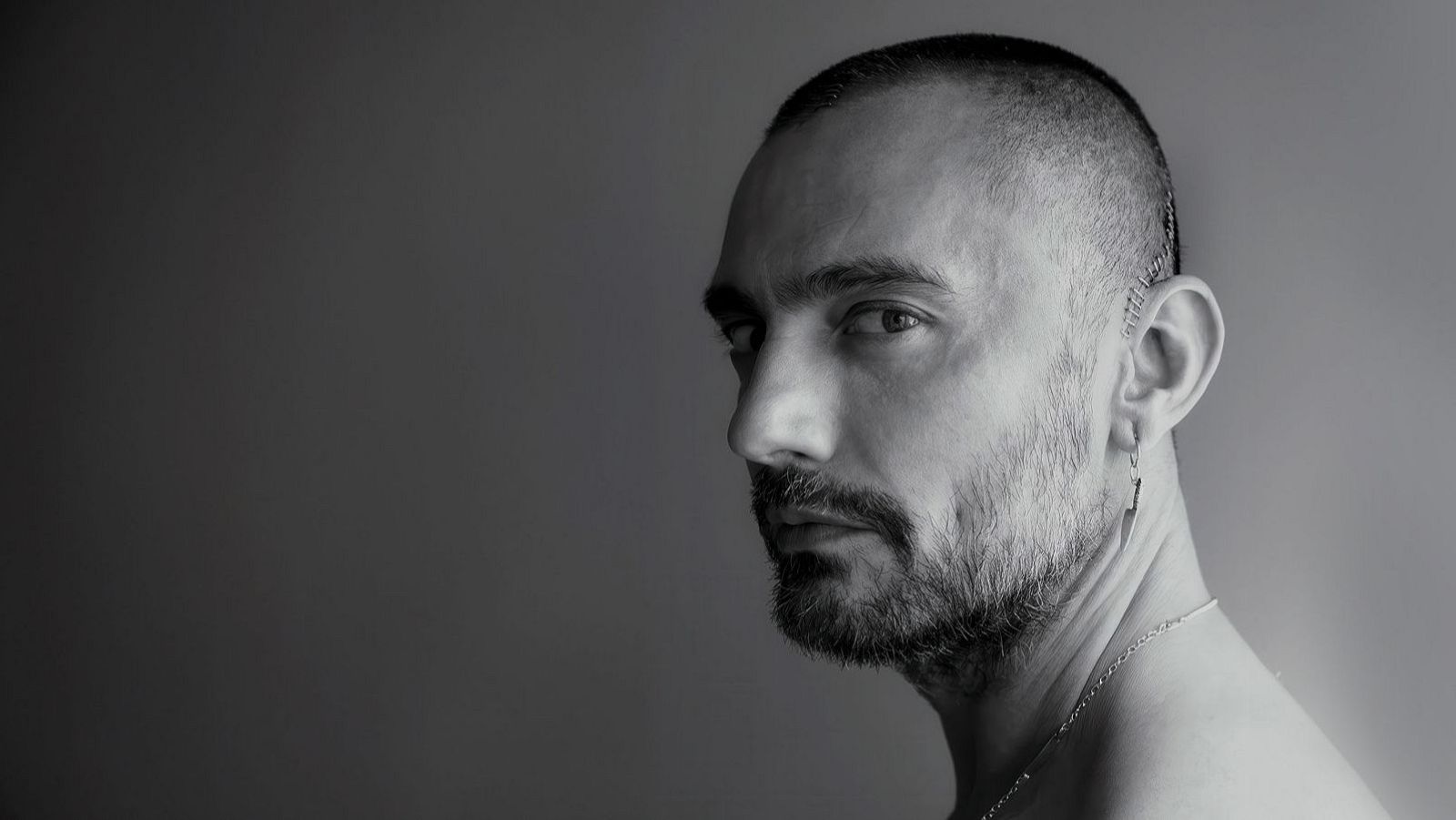 Retrato en blanco y negro de un hombre con cabello oscuro y corto, barba incipiente, pendiente de rayo y cadena, mirando hacia la derecha.