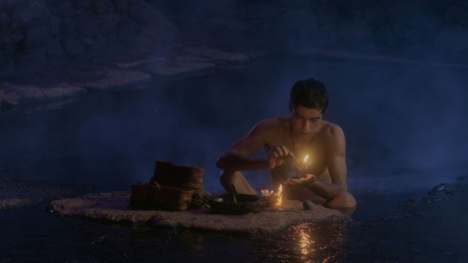 Un hombre joven de piel morena, con el torso desnudo, realiza un ritual en un entorno acuático oscuro, sosteniendo una llama y con un cuenco metálico frente a él. La luz del fuego ilumina la escena.