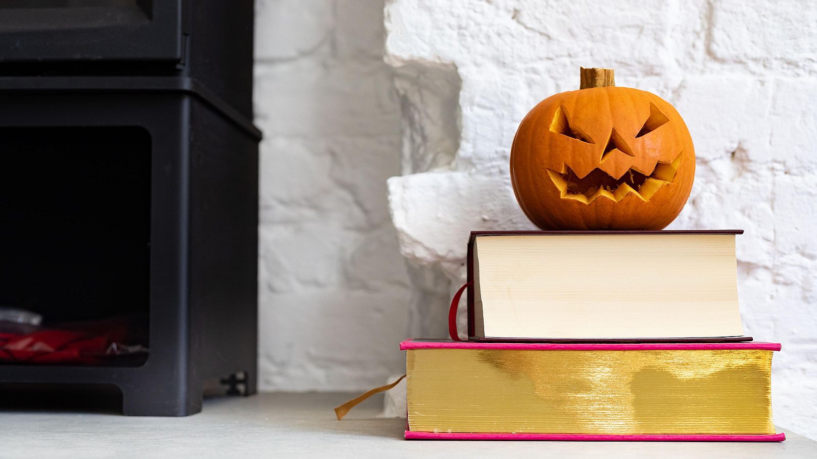 Una calabaza naranja tallada con una cara de Halloween descansa sobre una pila de libros. El libro inferior tiene una cubierta dorada y el superior es de color crema, todo sobre una superficie gris.