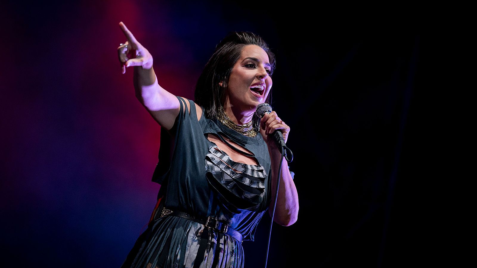 En un escenario oscuro iluminado por luces rojas y azules, una cantante con cabello oscuro y un vestido con detalles rasgados, sostiene un micrófono y canta con entusiasmo.