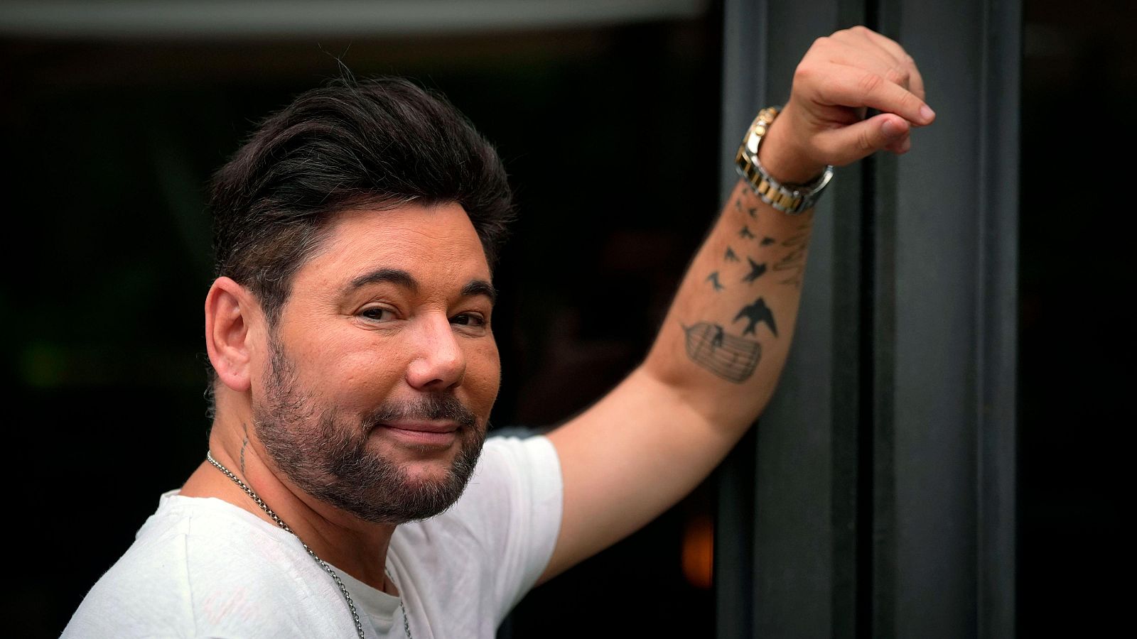 Miguel Poveda, con barba y cabello oscuro, luce una camiseta blanca y una cadena. Sus tatuajes, incluyendo una jaula con un pájaro, son visibles en su brazo, junto a un reloj dorado.