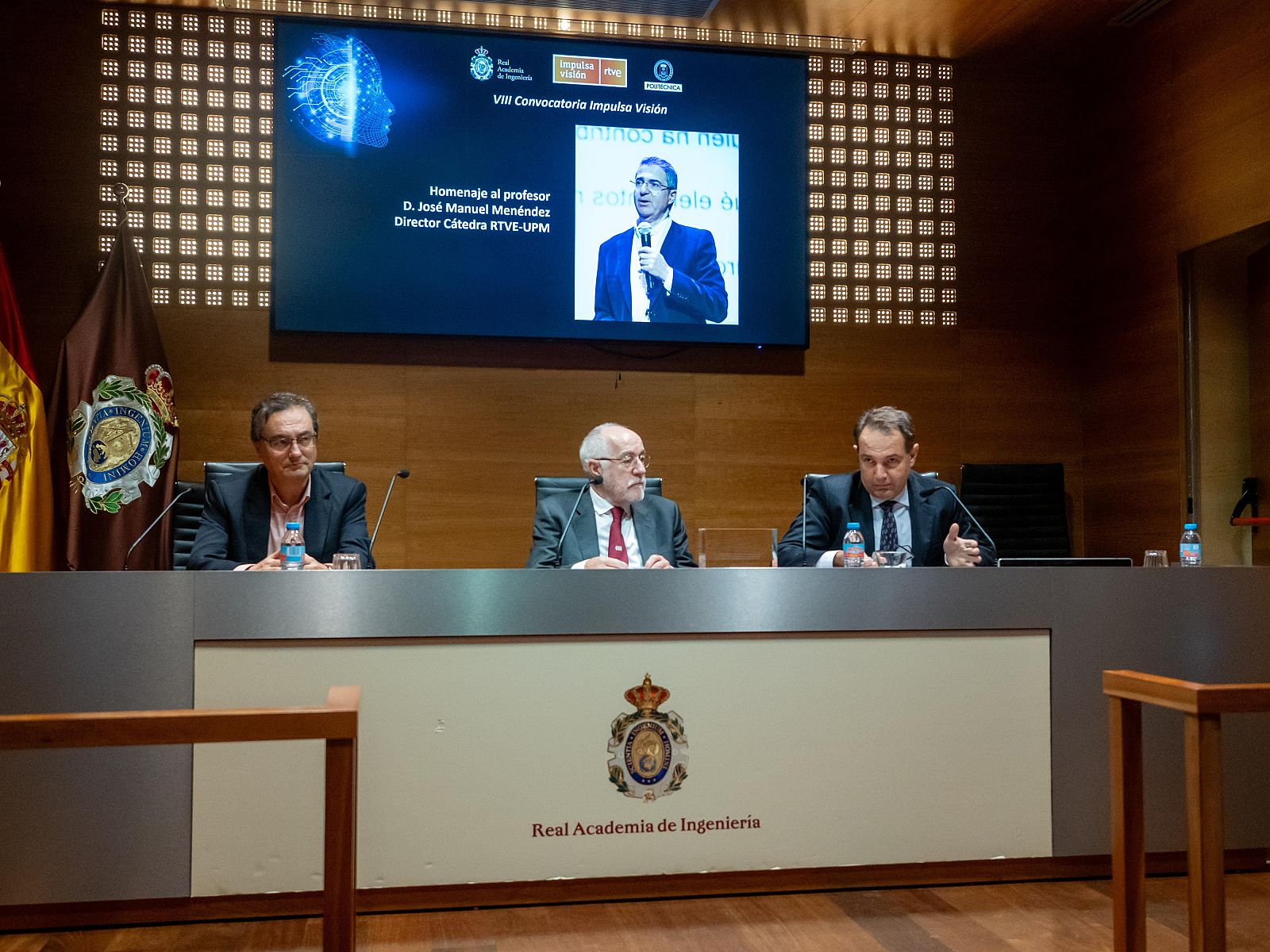En la imagen, tres hombres participan en un evento frente a una mesa con el logo de una institución. Detrás de ellos, una pantalla muestra un logo y un homenaje a una persona.
