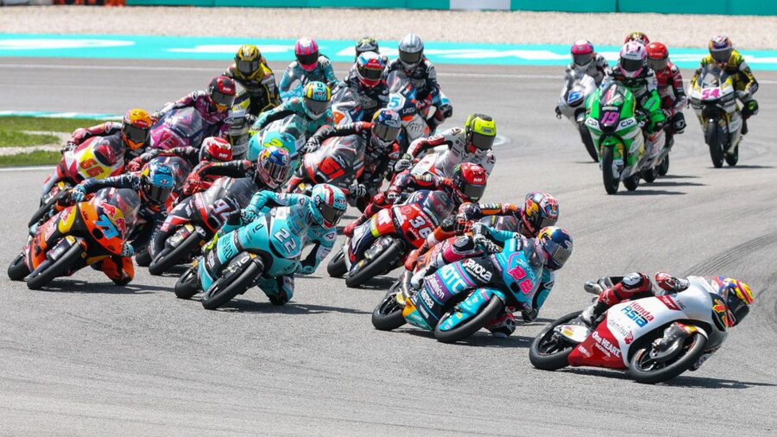En una pista de asfalto, varias motocicletas de distintos colores y diseños compiten en una carrera. Se distinguen números en las motos y los pilotos visten trajes y cascos protectores, con el cielo despejado como telón de fondo.