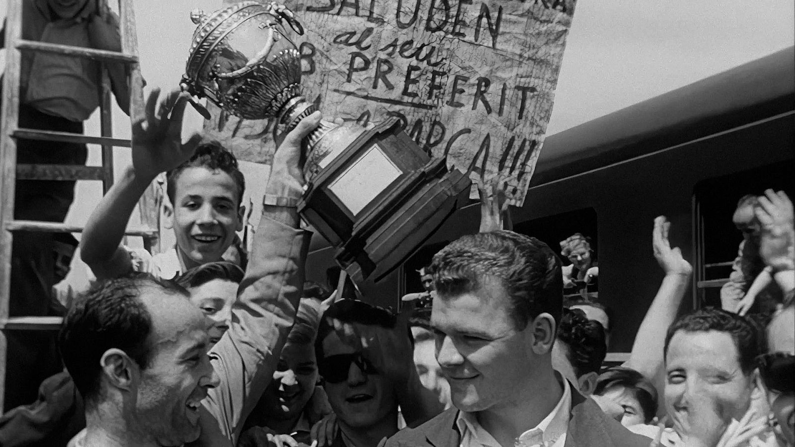 Aficionats celebrant un triomf esportiu al voltant d'un tren, amb una pancarta i una copa alçada. La imatge en blanc i negre reflecteix l'alegria i l'entusiasme d'un esdeveniment victoriós.