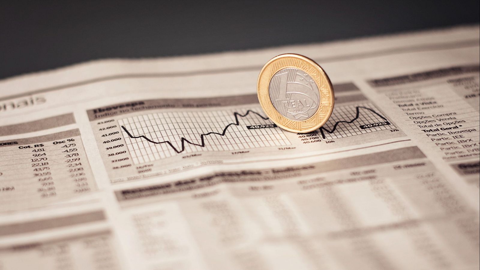 Un periódico económico abierto muestra un gráfico de líneas del índice Ibovespa y una moneda de 1 real brasileño. Se observan tablas con datos financieros y términos como "Opções Venda" y "Partic. Ibovespa".