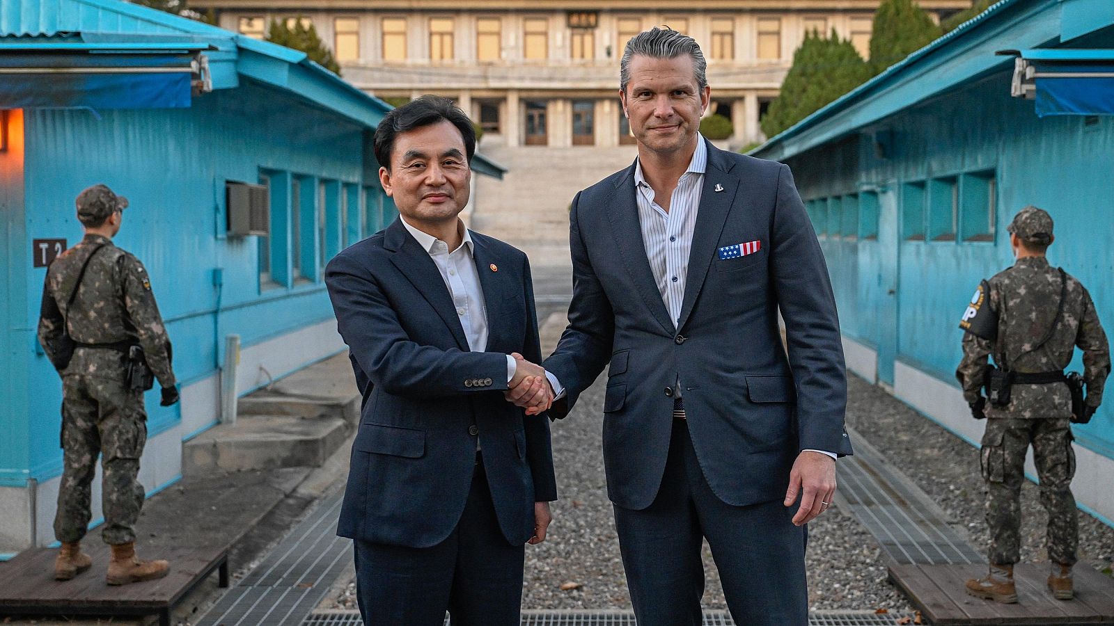 Dos hombres, uno de ellos el homólogo surcoreano, se estrechan la mano frente a una construcción azul clara, flanqueados por militares uniformados. Se observa un edificio de varios pisos en el fondo.