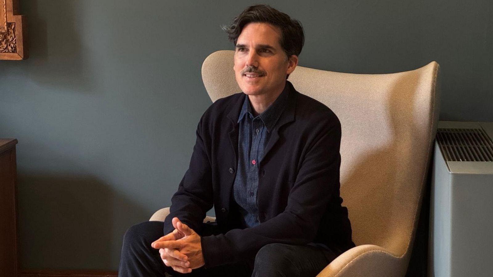 Pablo Maurette, con camisa vaquera y chaqueta oscura, está sentado en un sillón claro, con las manos entrelazadas. El fondo muestra una pared verde oscuro y un mueble de madera.