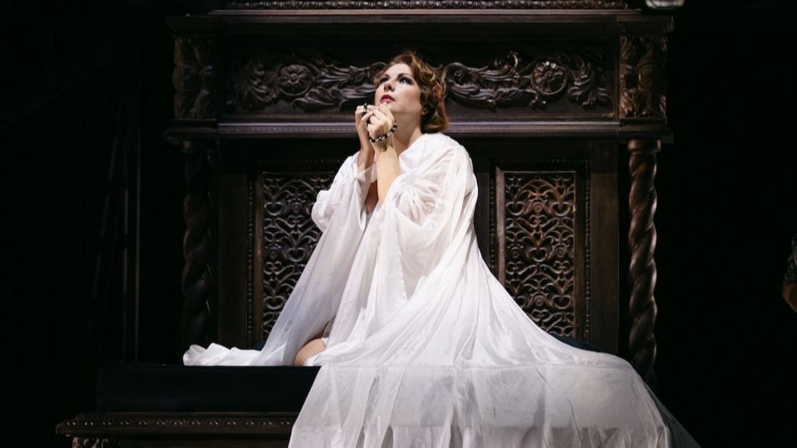 En una habitación con luz tenue, una mujer con camisón blanco yace sentada en una cama con cabecero de madera, con las manos juntas y la mirada dirigida hacia arriba.