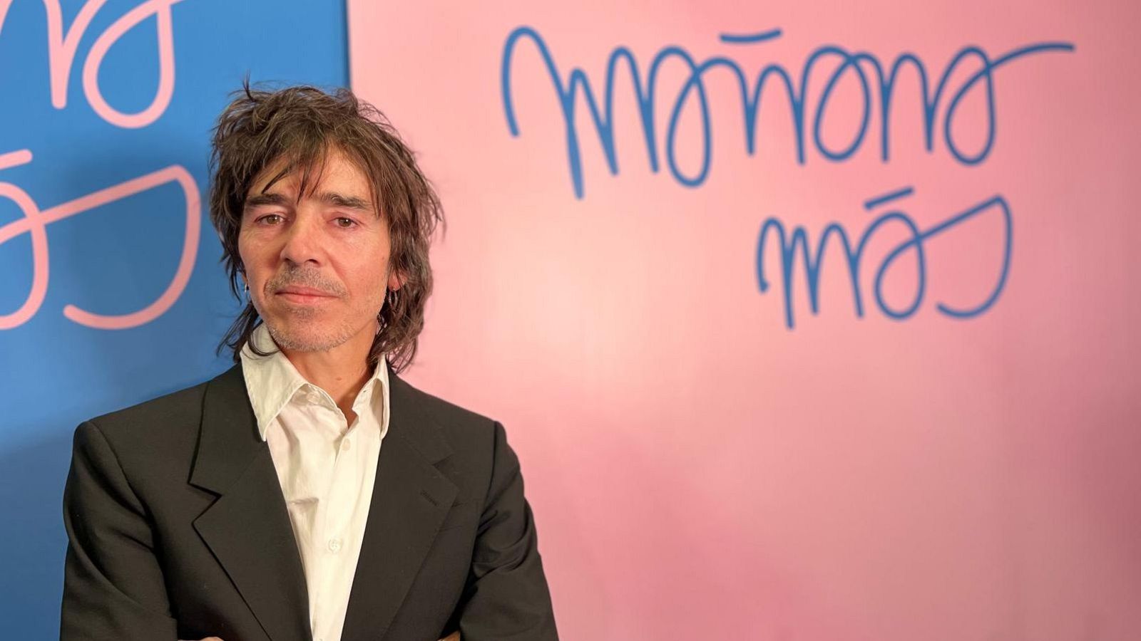 Rubén Pozo, con chaqueta negra y camisa blanca, posa con los brazos cruzados frente a un fondo dividido en azul y rosa, donde se lee una frase en azul.