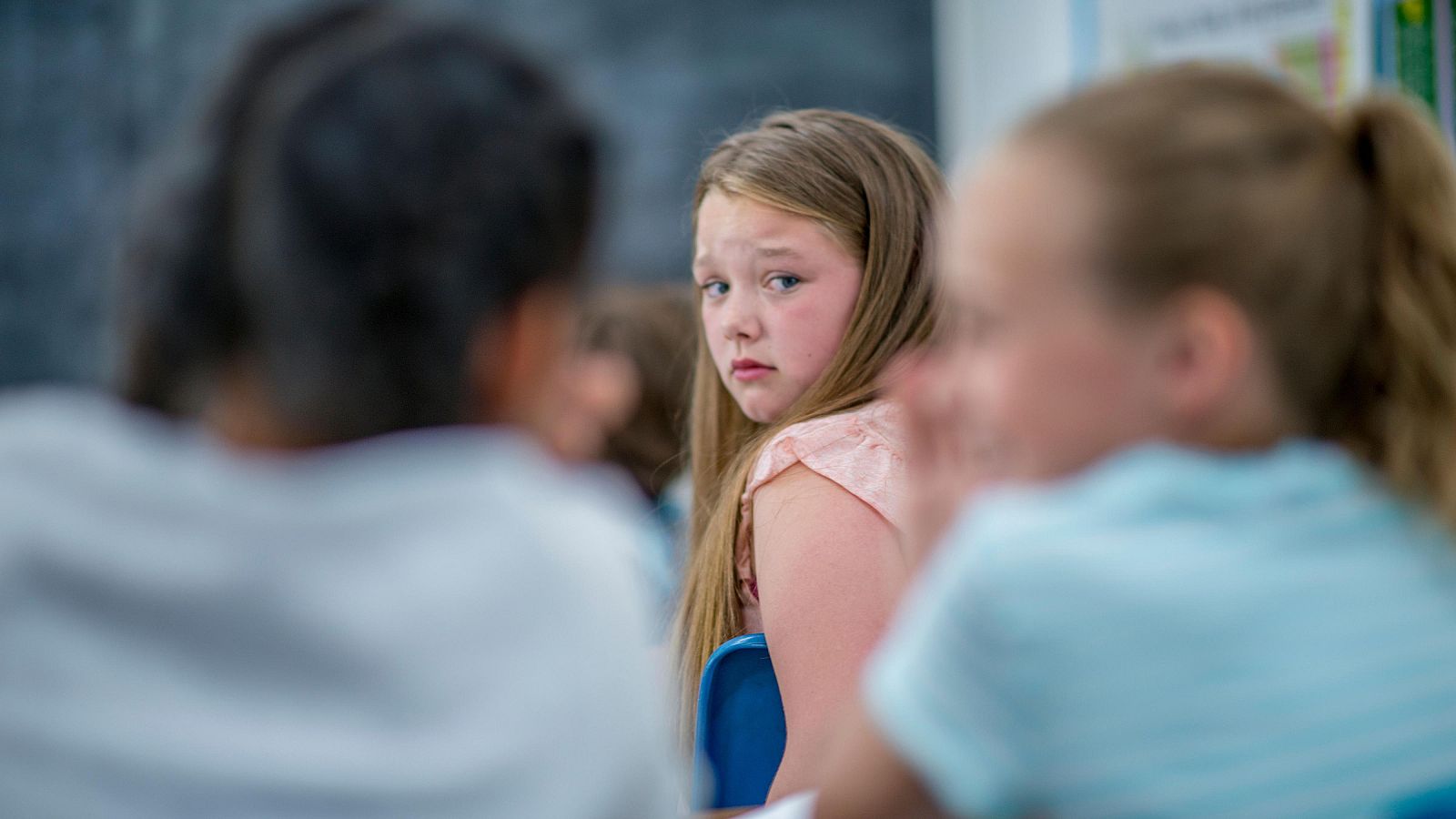 En un aula escolar, una niña rubia con ojos azules y blusa rosa, muestra una expresión de tristeza mientras mira hacia atrás. Otros niños están presentes, algunos parecen estar cuchicheando, sugiriendo un posible ambiente de acoso.