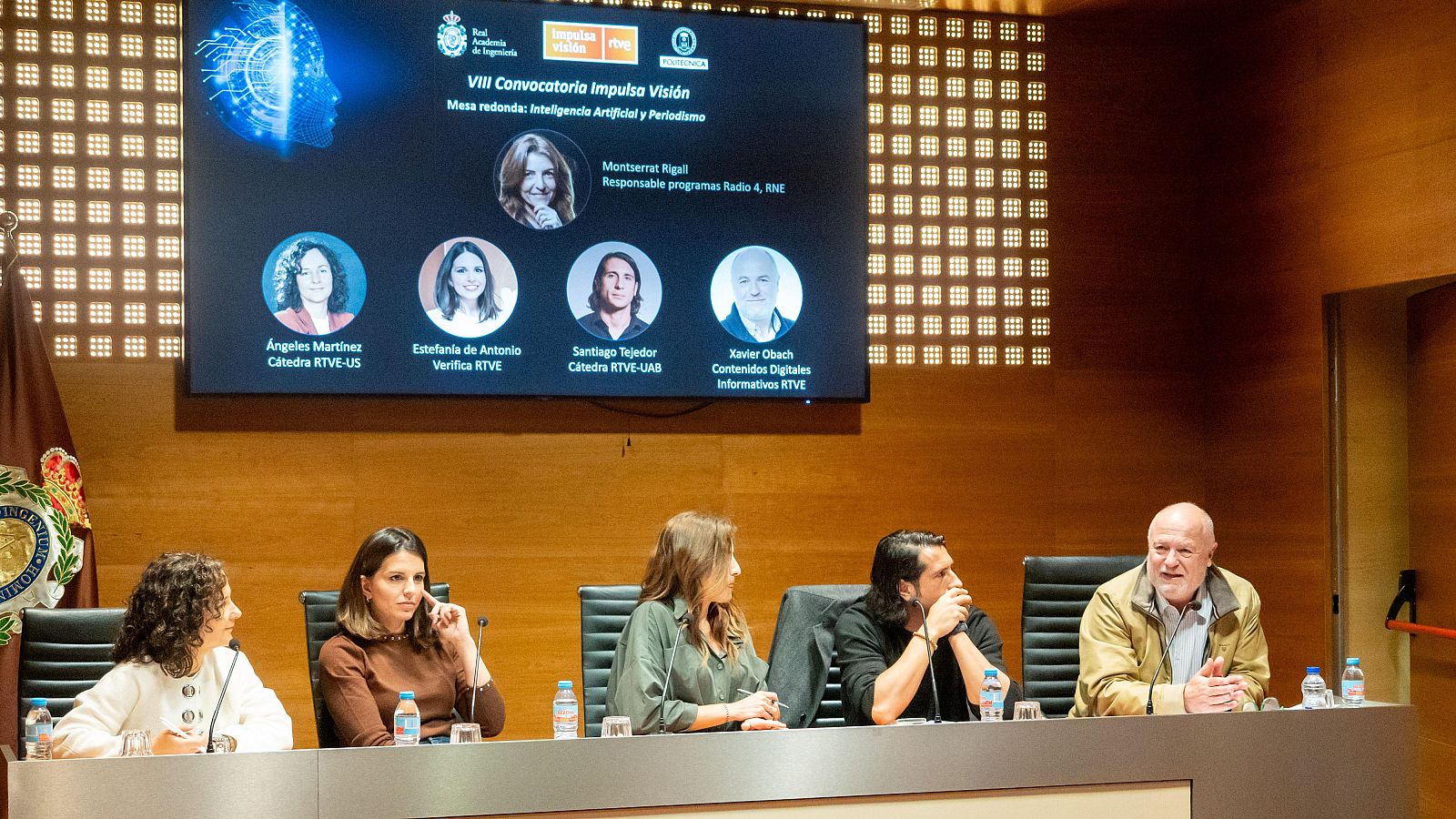 Una mesa redonda con cinco participantes discute sobre Inteligencia Artificial y Periodismo. En la parte superior, una pantalla muestra el título del evento y los nombres de los ponentes, mientras que en la mesa hay botellas de agua.