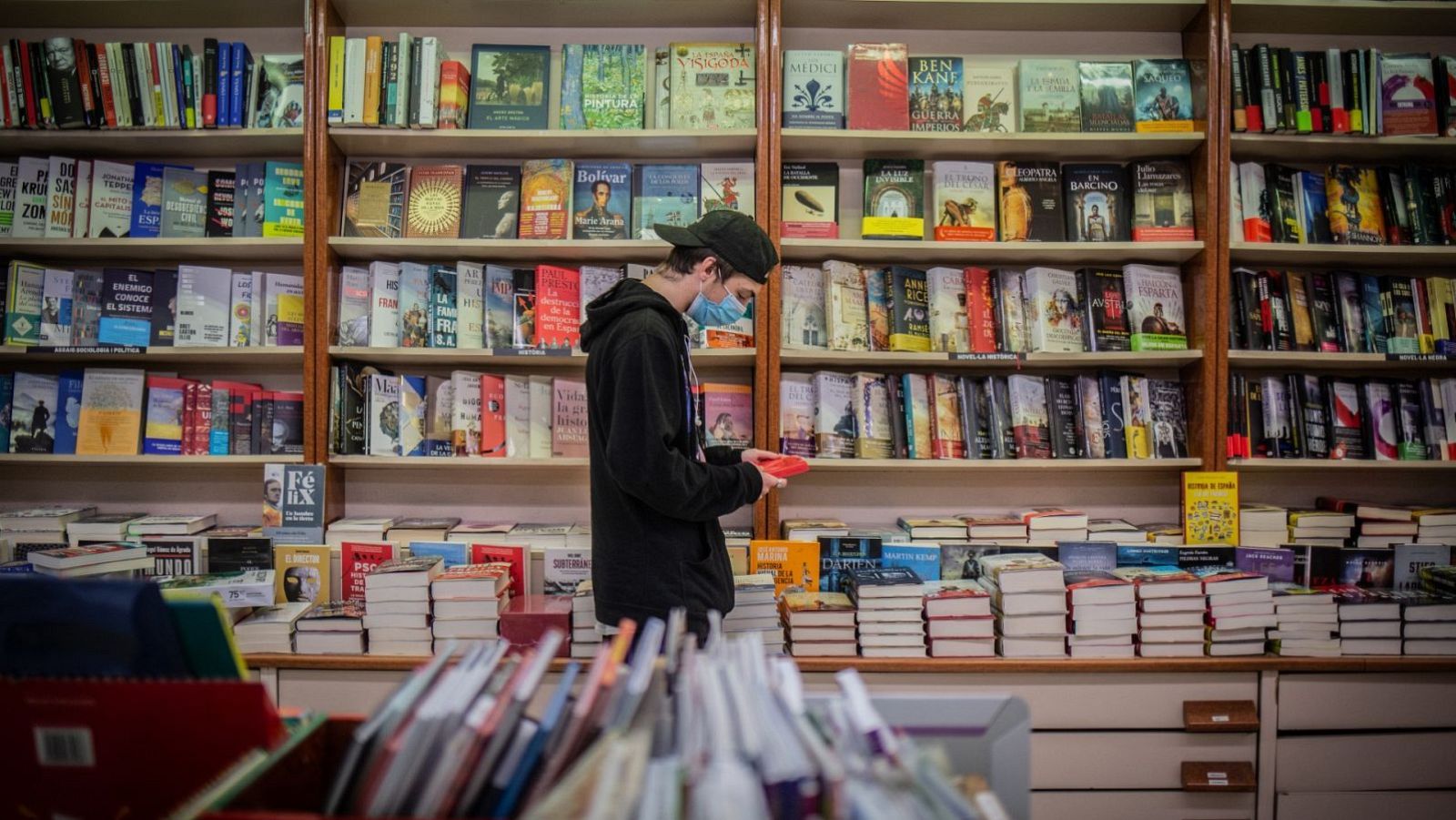 Un joven con mascarilla y gorra, de pie frente a una estantería repleta de libros, sostiene un libro rojo y lo examina en el interior de una librería.