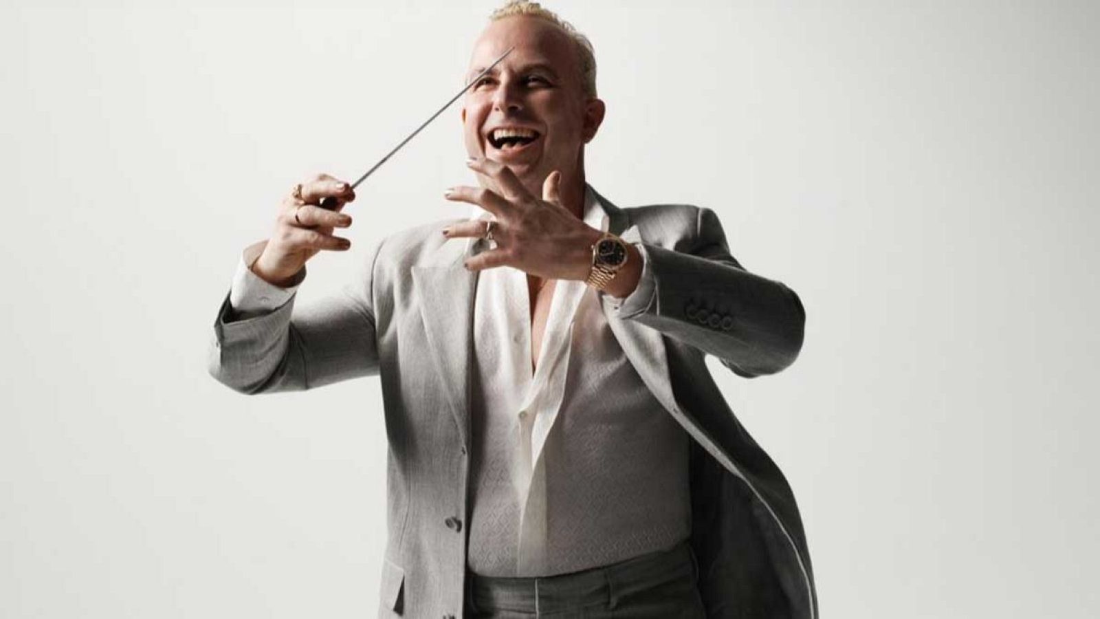 Yannick Nézet-Séguin, con traje gris claro y camisa blanca, dirige una orquesta con una batuta en la mano derecha, mostrando una sonrisa. Luce anillos y un reloj dorado.
