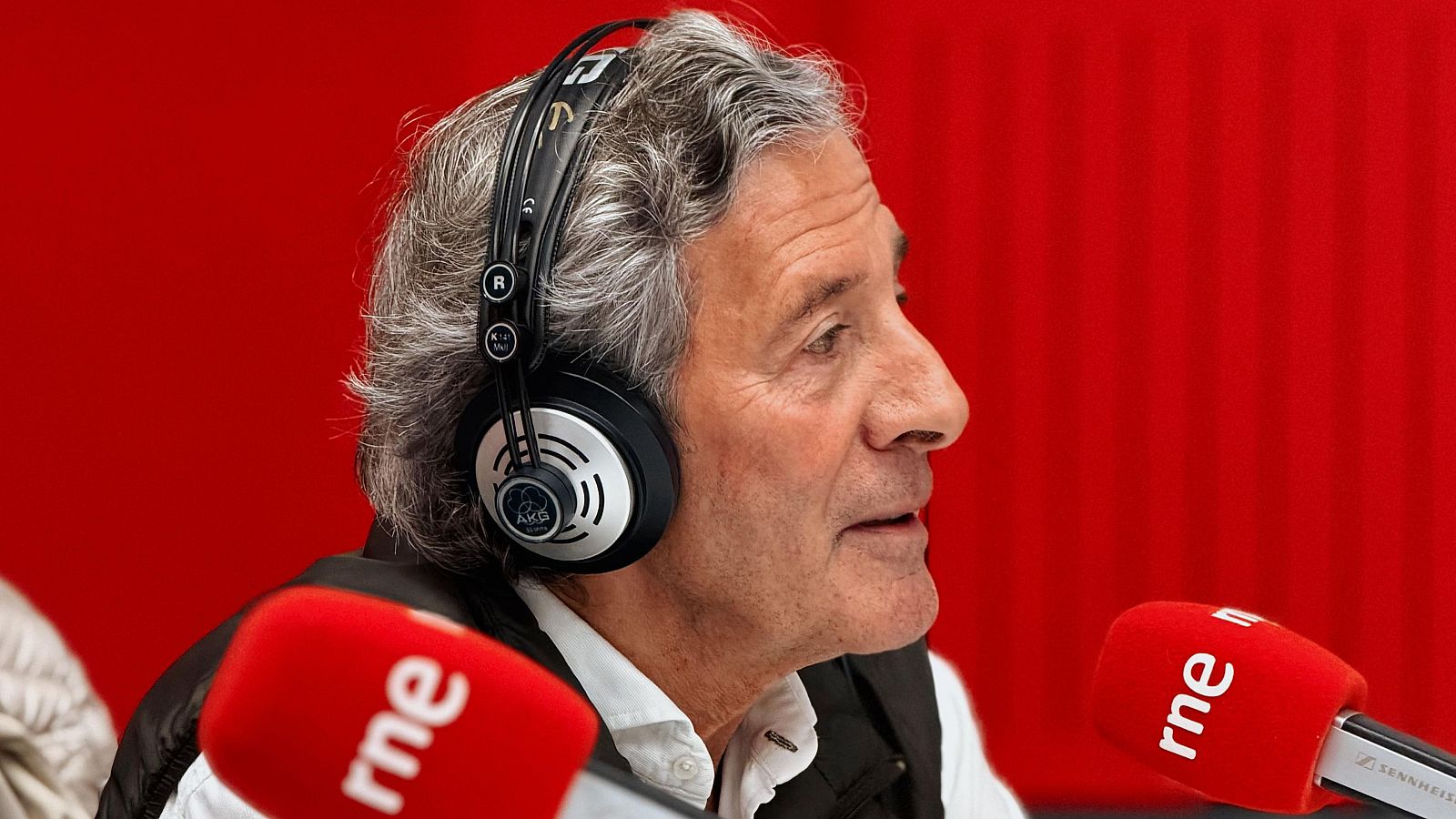 Jesús Álvarez, con auriculares y chaleco, se encuentra frente a dos micrófonos rojos de RNE, con un fondo rojo con líneas verticales.