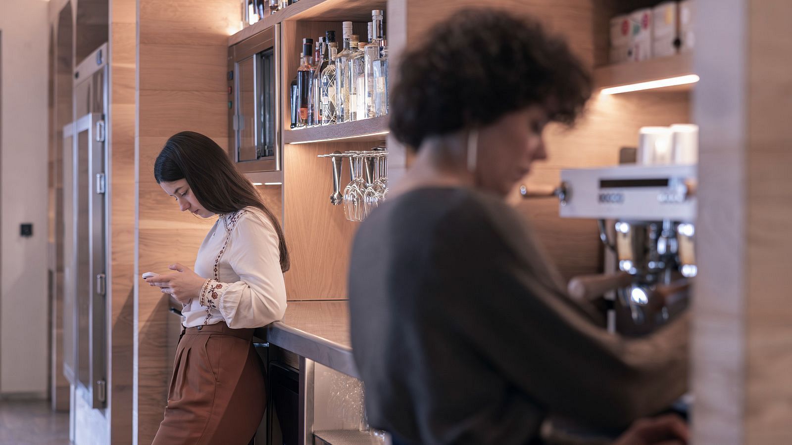 Dos mujeres interactúan en un bar: una joven revisa su teléfono mientras está apoyada en la barra, y otra prepara café tras la máquina, ambas en un entorno con decoración de madera y botellas.