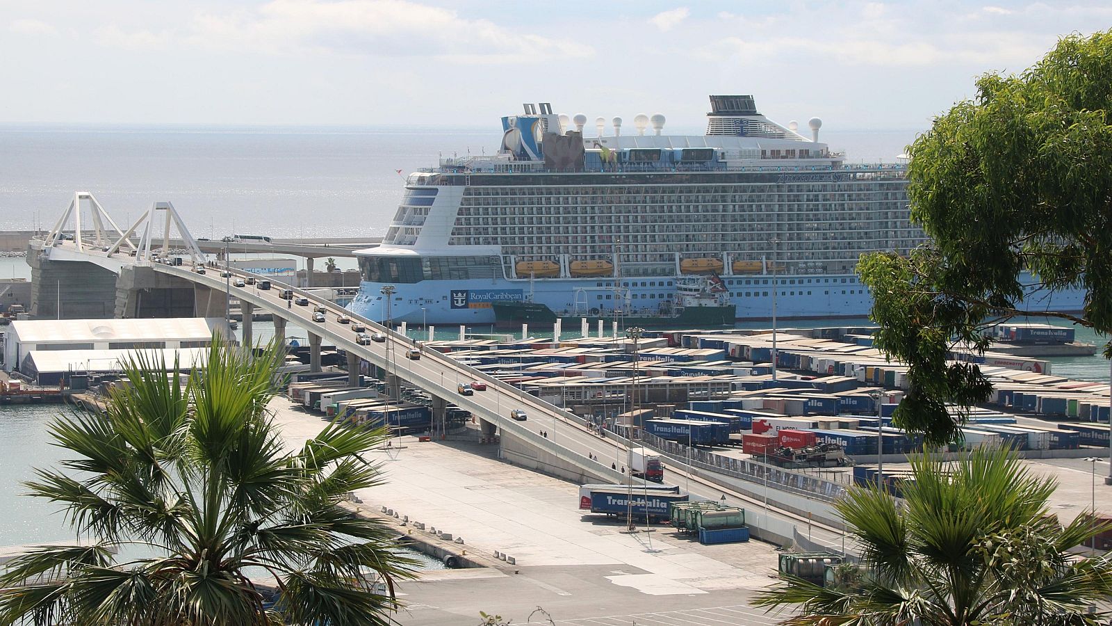 Un creuer blau i blanc de grans dimensions, amb múltiples cobertes i finestres, atracat al port, amb un pont i contenidors en primer pla.