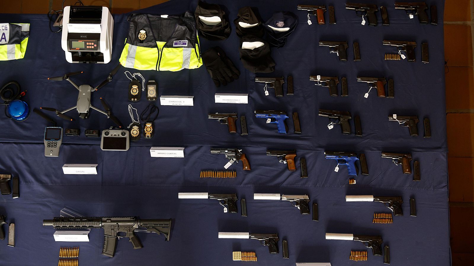 La fotografía muestra una variedad de objetos incautados, incluyendo un chaleco reflectante, una máquina contadora de billetes, un dron, armas de fuego, munición, y otros accesorios, posiblemente relacionados con una operación policial.