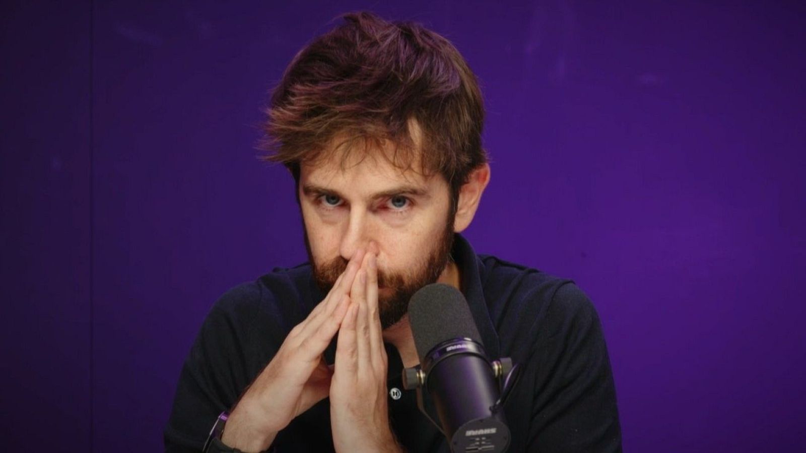 Miguel Campos, con barba incipiente y camisa oscura, aparece pensativo frente a un micrófono negro, con las manos entrelazadas. El fondo es de color morado.