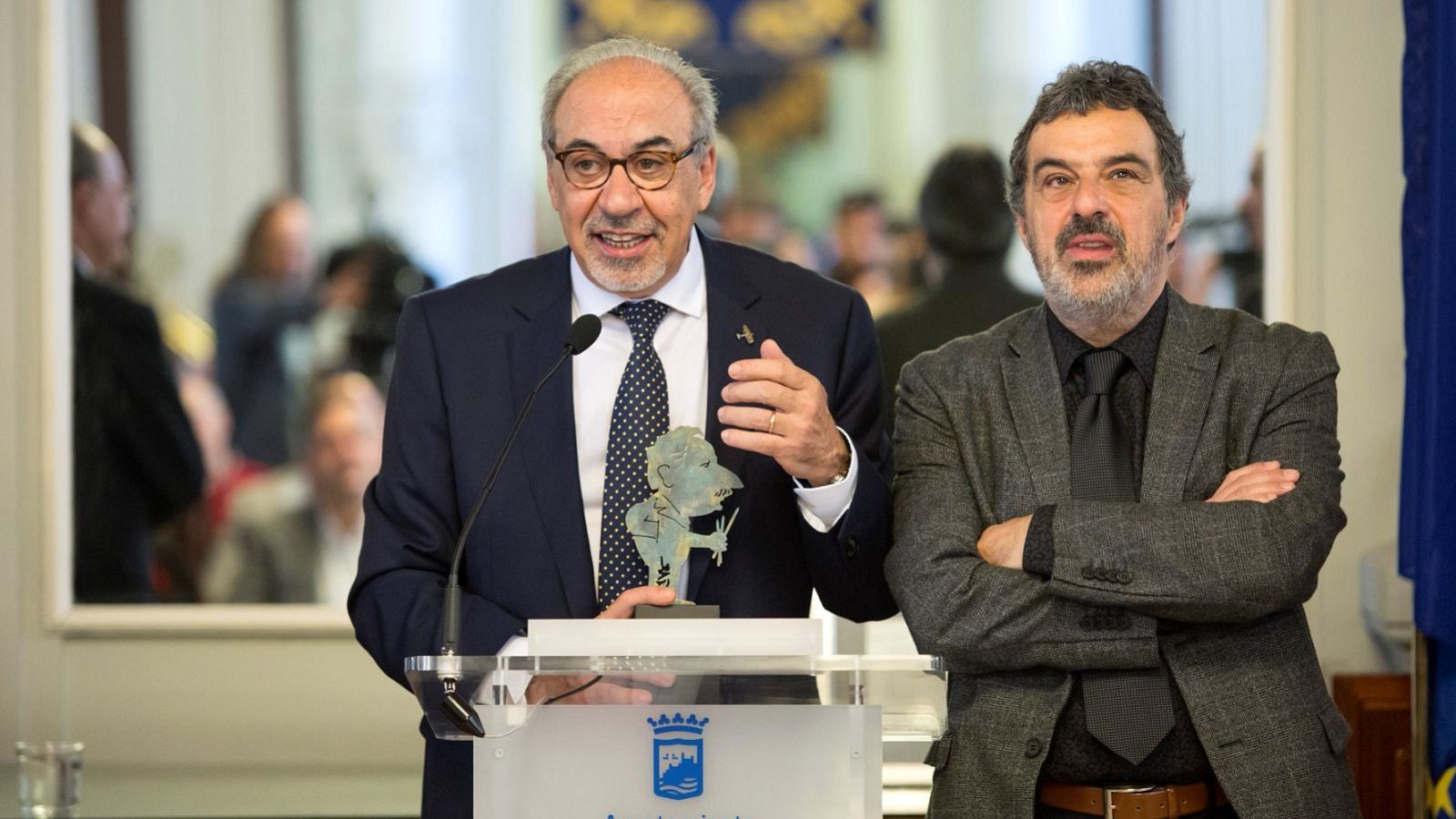Dos hombres, José María Gallego y Julián Rey, posan en un evento. Uno sonríe con una estatuilla, mientras el otro, con los brazos cruzados, muestra una expresión seria.