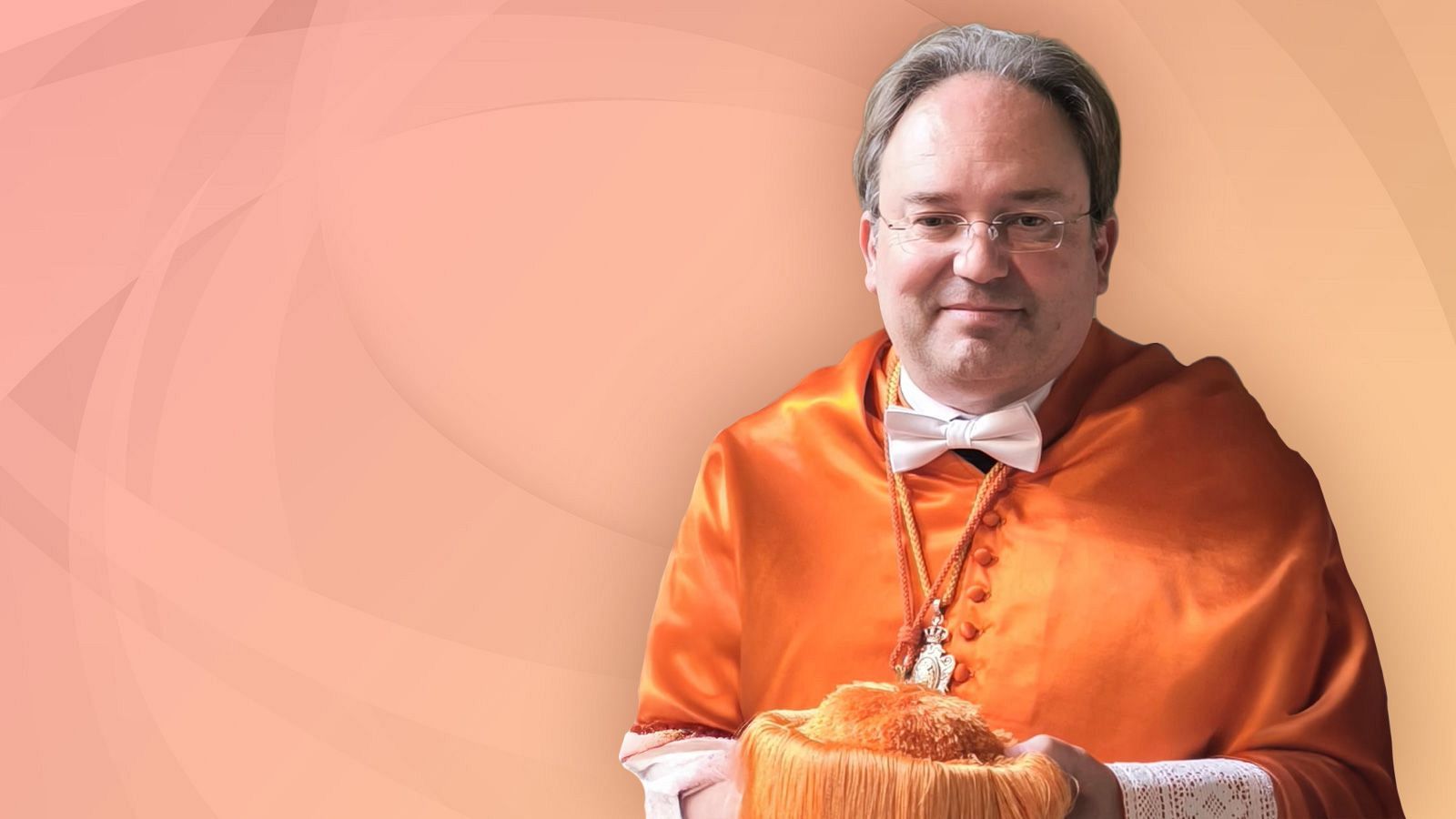 Félix Ortega, con toga y birrete, posa para la cámara. Destacan sus gafas, pajarita y un collar con colgante. La imagen incluye su nombre y la mención de "Cátedras RTVE".