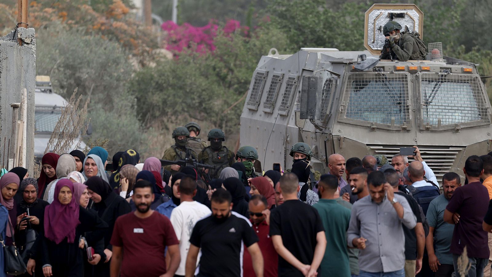 En un campo de refugiados, un grupo de civiles se enfrenta a soldados armados. Un vehículo blindado se encuentra en la escena, con un soldado apuntando un arma. El ambiente refleja tensión y preocupación.