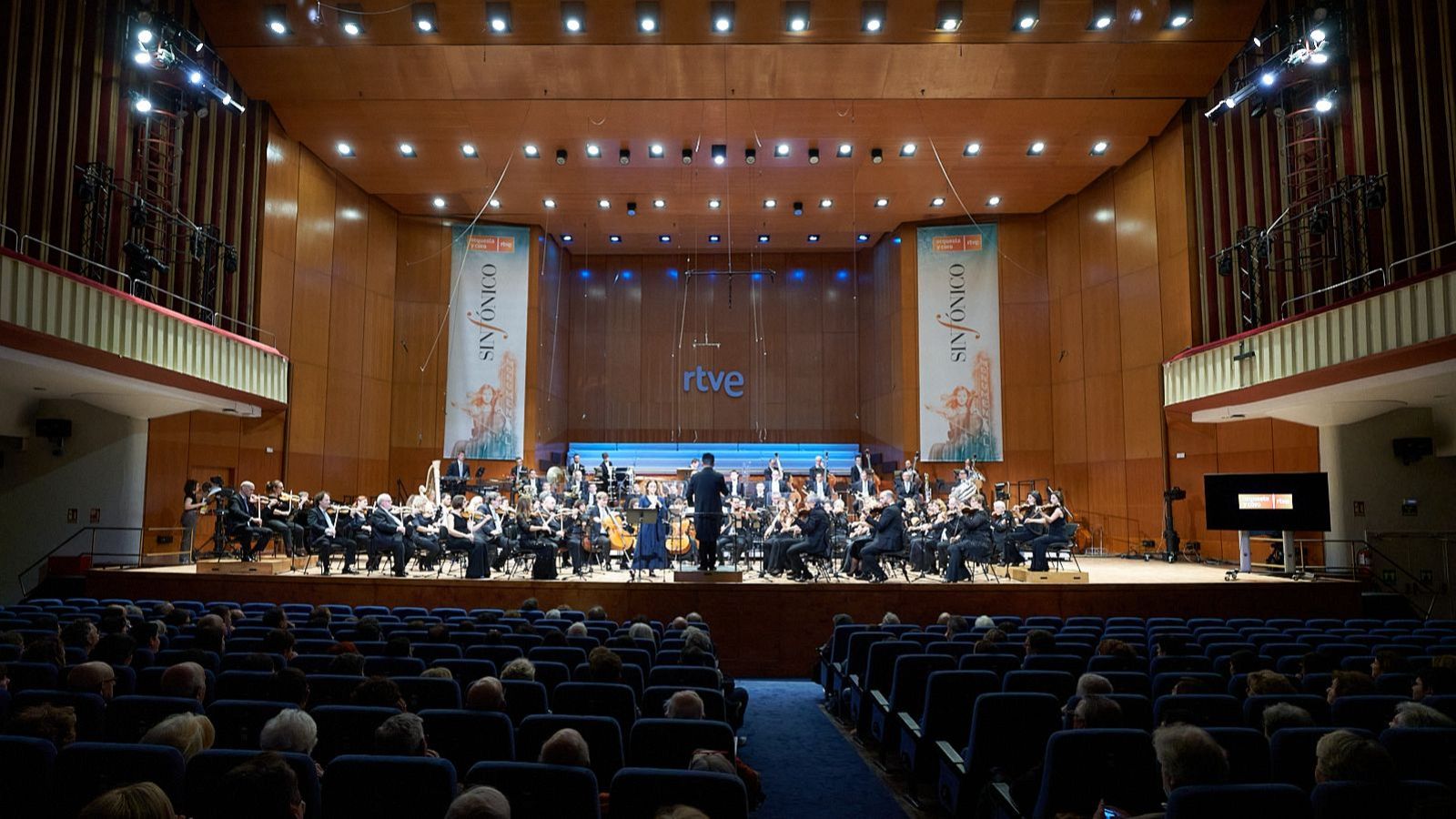 Una orquesta completa, con un director de orquesta de espaldas, ofrece un concierto en un auditorio lleno. Se observan instrumentos musicales y el público disfruta del espectáculo, con banners laterales que anuncian el evento.
