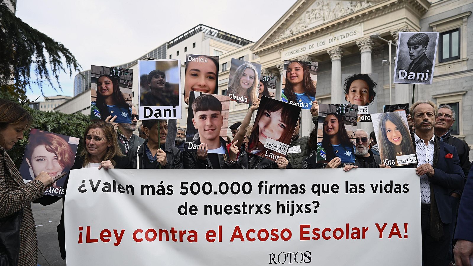 Manifestación frente al Congreso de los Diputados con pancartas que muestran fotografías y nombres de jóvenes, exigiendo una ley contra el acoso escolar. Destaca una pancarta central con un mensaje sobre la importancia de las vidas de los afectados.