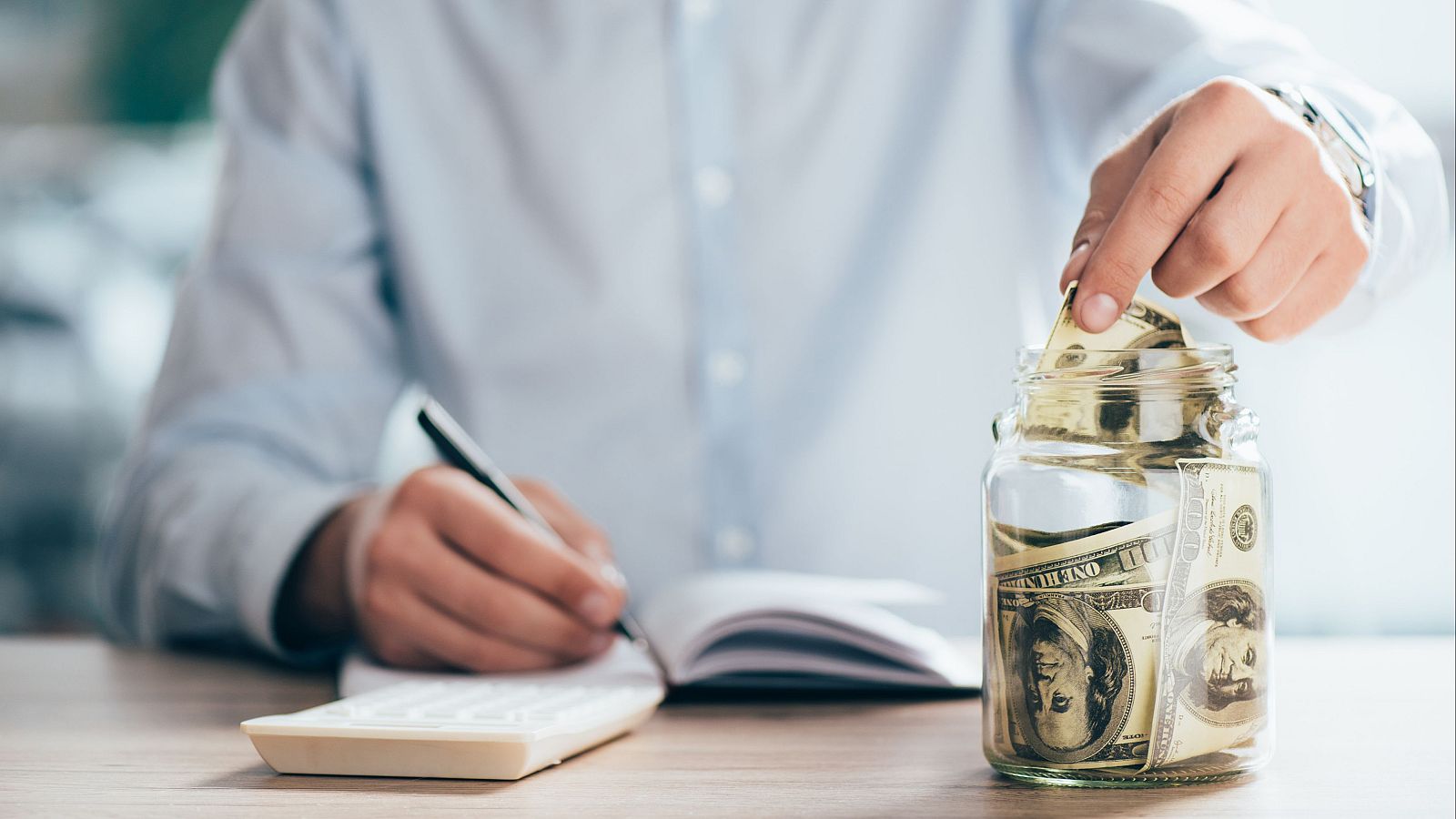 En la imagen, una persona inserta un billete en un recipiente lleno de dinero, con una calculadora, un bolígrafo y una libreta cerca, sobre una mesa de madera, posiblemente en un contexto de ahorro.