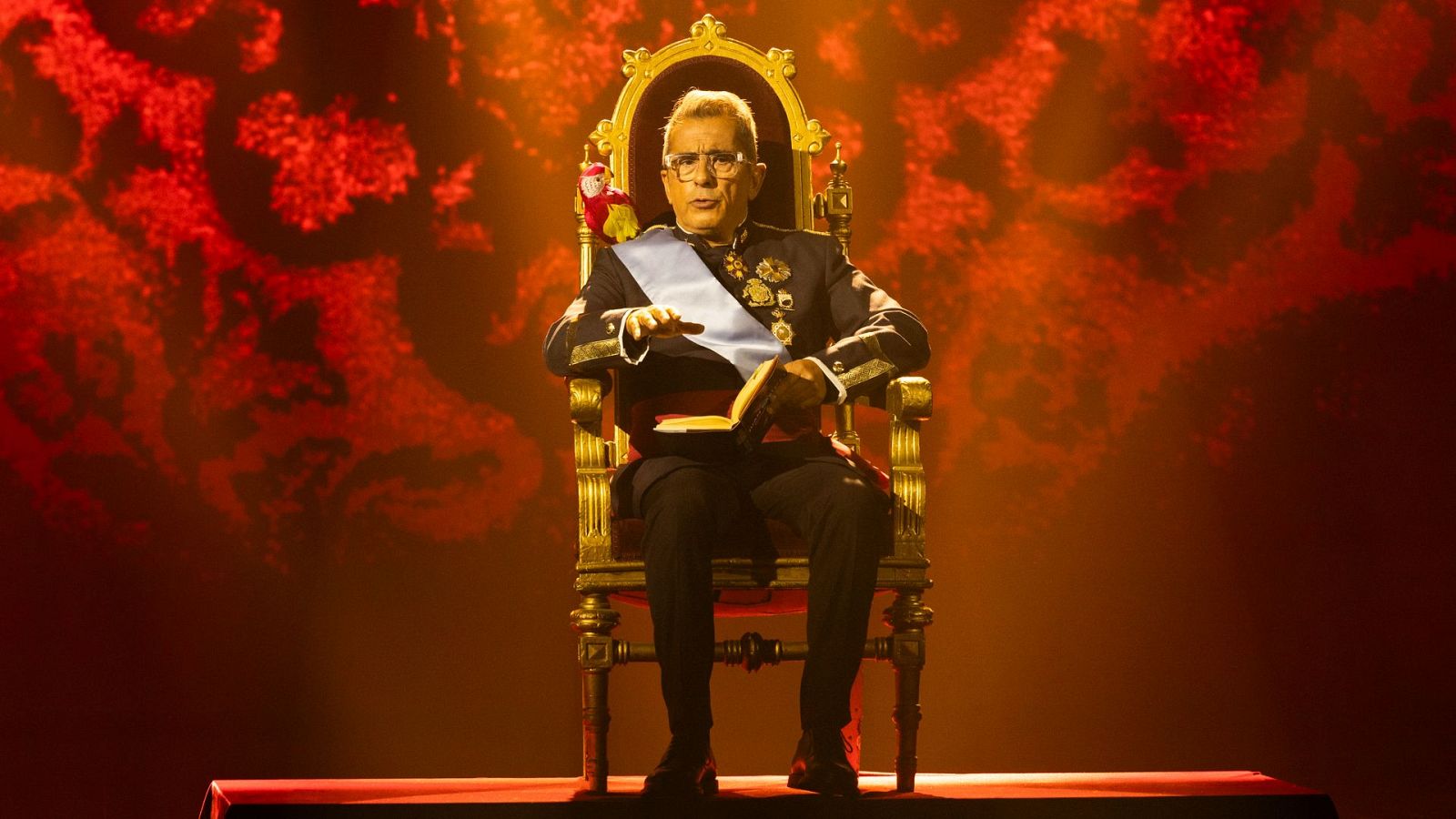 Andreu Buenafuente, con gafas y traje regio, posa sentado en un trono dorado con un libro en las manos, con un loro de peluche a su lado, sobre un fondo rojo.