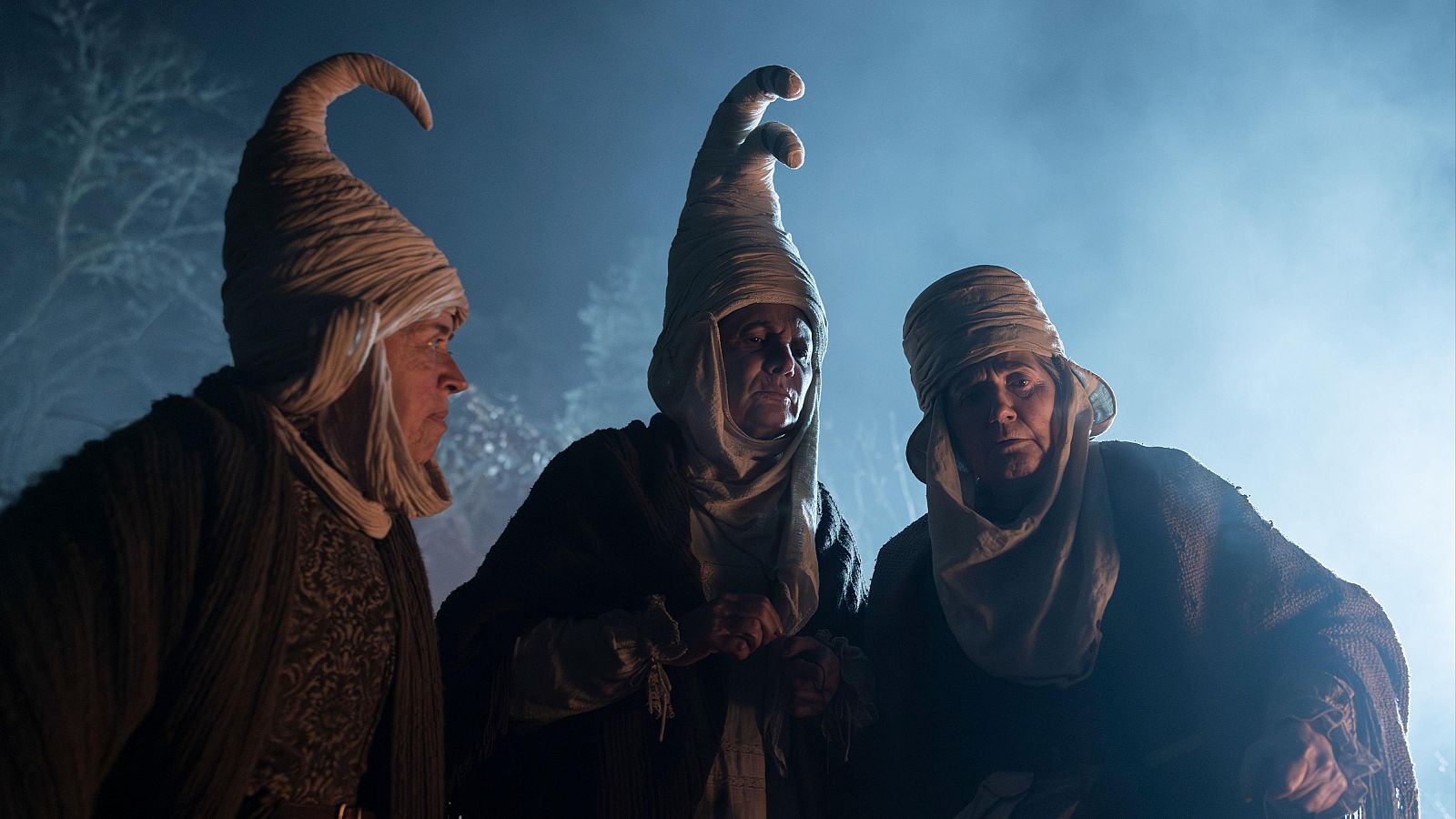Un fotograma de la película muestra a tres figuras con sombreros altos y oscuros en un ambiente nocturno. La iluminación tenue desde abajo crea un ambiente misterioso y sugiere un contexto histórico o folclórico.