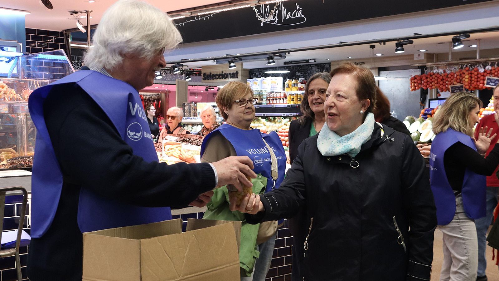 En un supermercat, un voluntari amb peto blau entrega una bossa a una dona. Altres persones, algunes també amb petos, es troben a la botiga, amb prestatgeries i un mostrador de carnisseria al fons.