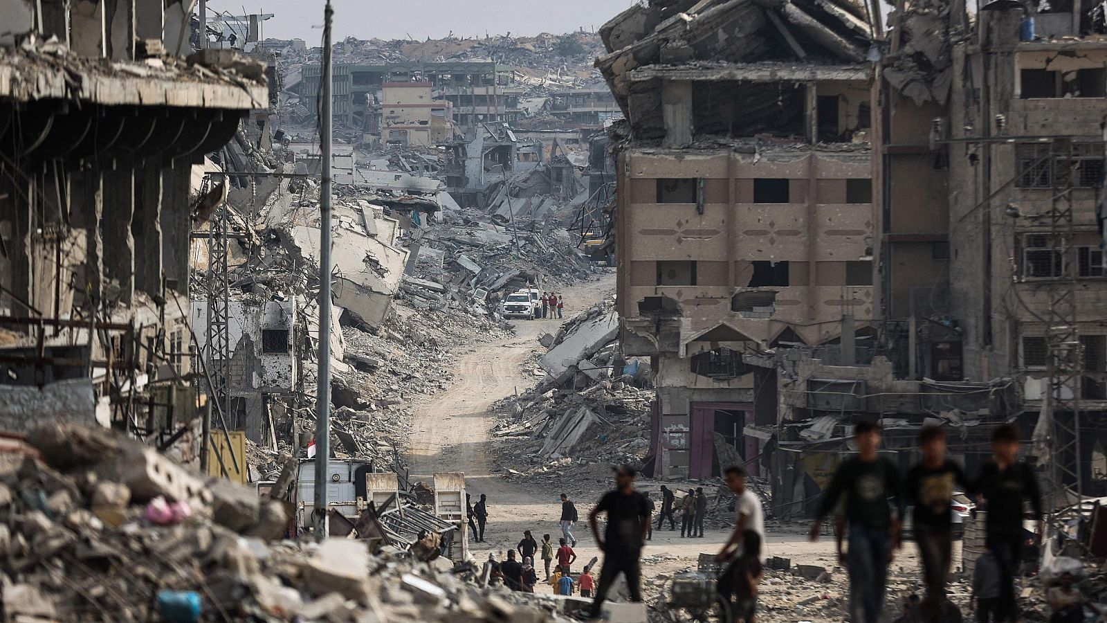 Una escena de devastación urbana muestra edificios colapsados y dañados, con escombros esparcidos. Personas caminan entre las ruinas, mientras que un vehículo se observa a lo lejos, transmitiendo una sensación de desolación.
