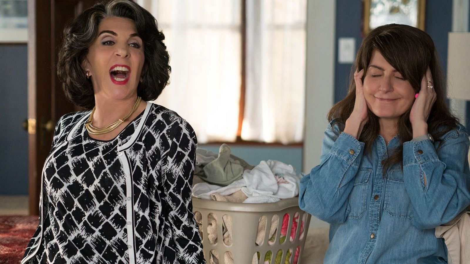 Dos actrices, una con blusa estampada y otra con camisa vaquera, interactúan cerca de una cesta de ropa. Una habla con entusiasmo, la otra se tapa las orejas.