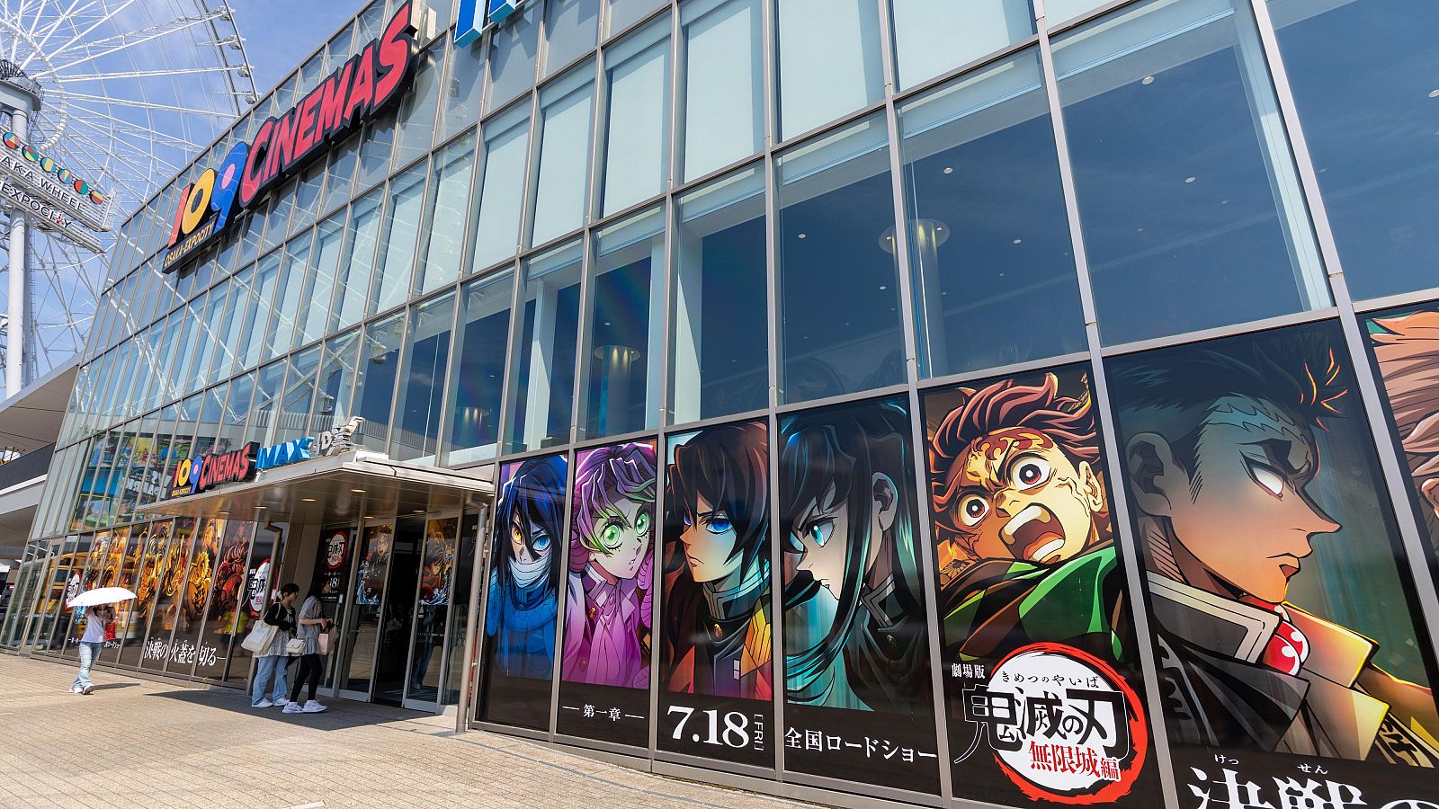 Un cine en Osaka, Japón, presenta carteles de una película a través de grandes ventanales. Se ven personas caminando y entrando al edificio, con una noria al fondo.