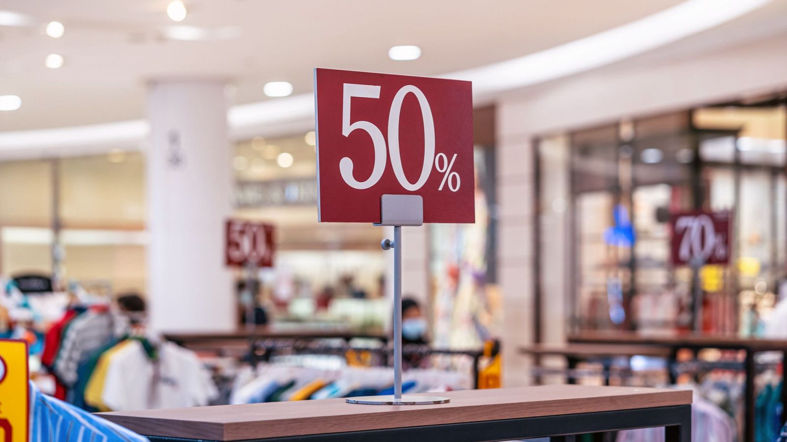 Cartel rojo con texto blanco anuncia un 50% de descuento en rebajas. Se observa en una mesa de madera oscura, con otros carteles de rebajas en el fondo.