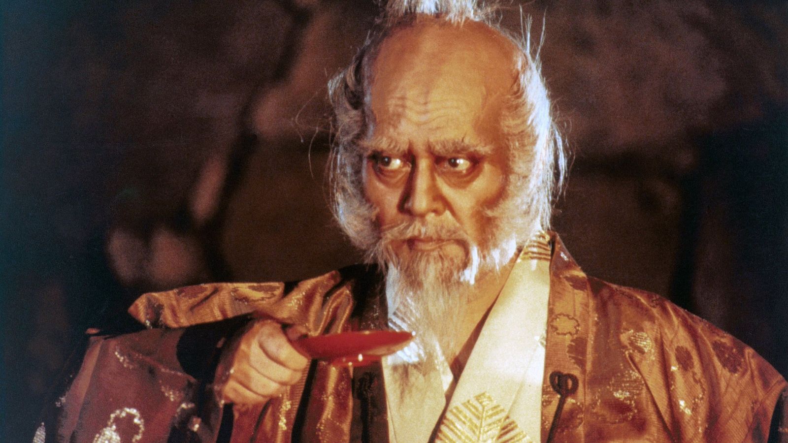 En la imagen, un hombre con cabello blanco recogido y barba prominente, vestido con un kimono estampado, sostiene una copa. Su mirada es intensa.