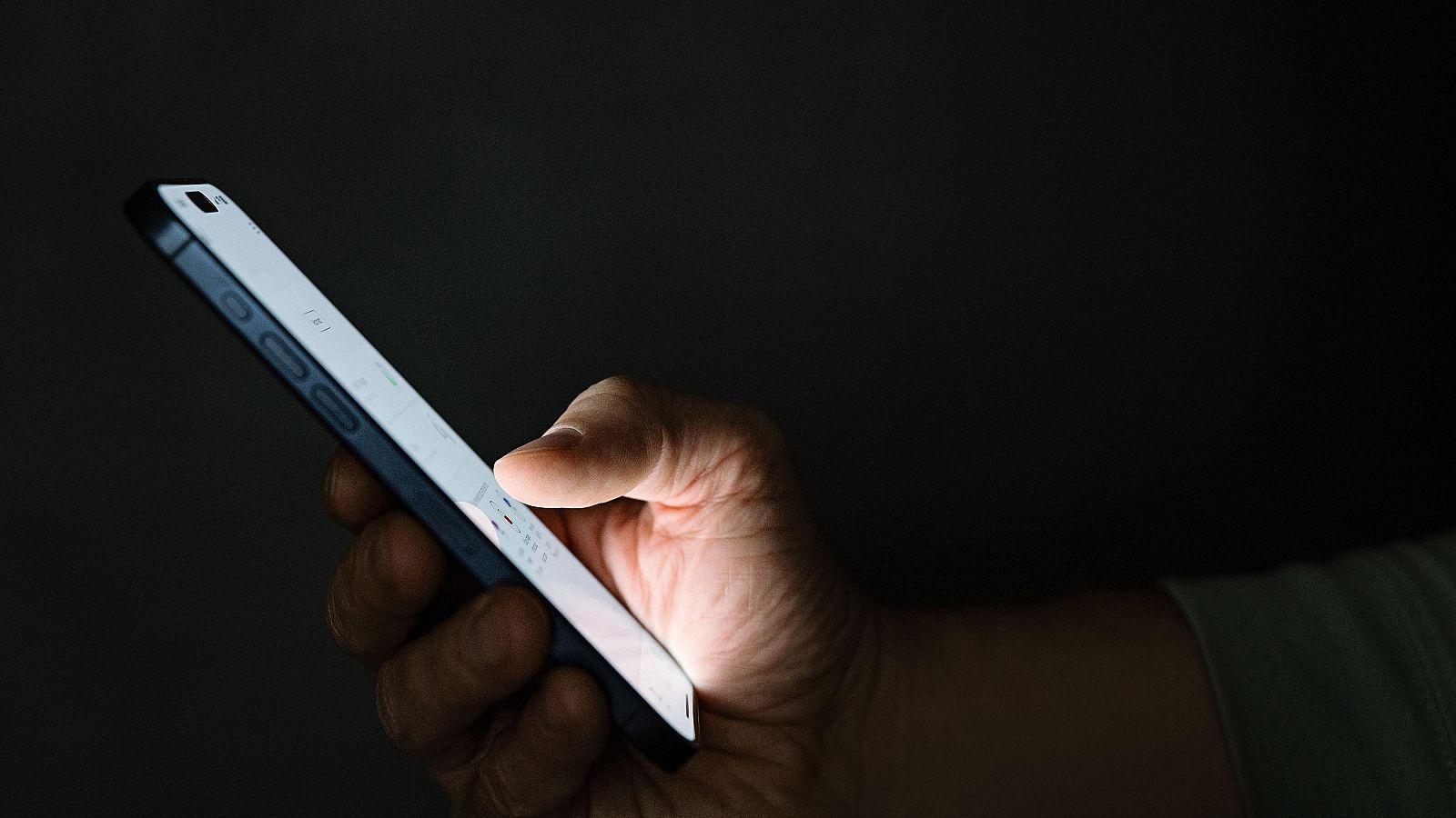Una mano interactúa con la pantalla iluminada de un teléfono móvil, posiblemente navegando o utilizando alguna aplicación. El fondo oscuro centra la atención en el dispositivo.