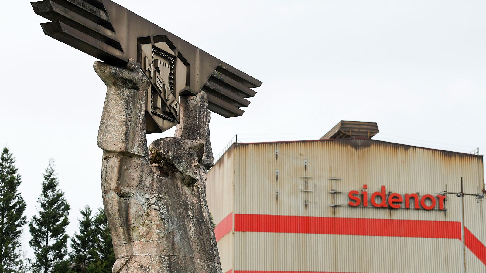 Una escultura con el logo de Sidenor se alza frente a un edificio industrial blanco con franjas rojas y el nombre de la empresa. El cielo gris y árboles verdes enmarcan la escena.