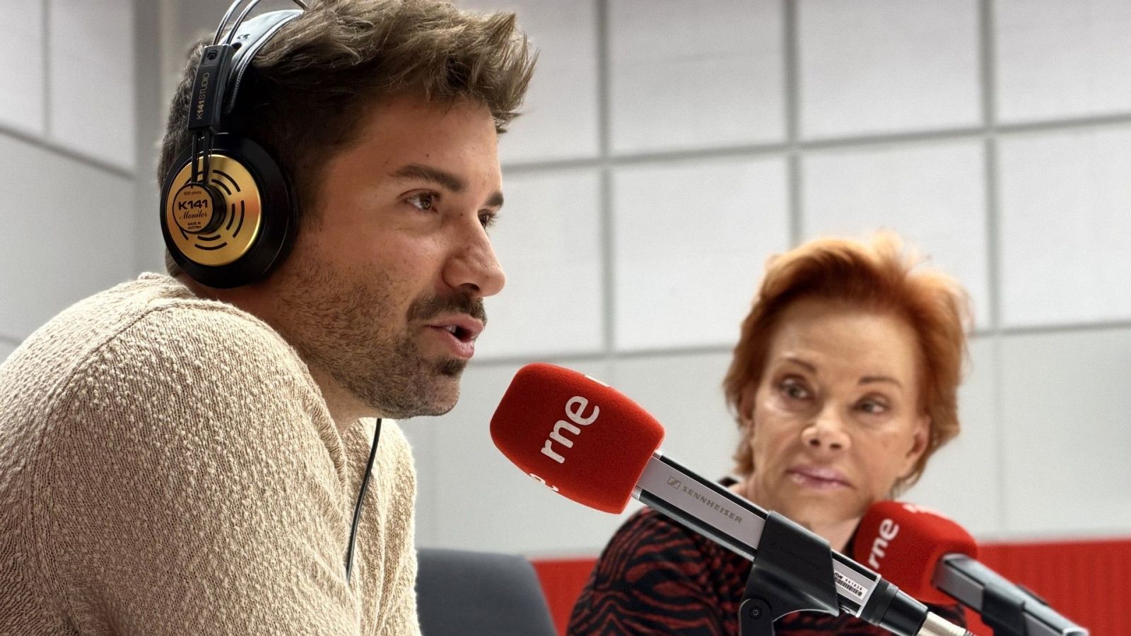 Pablo Alborán y Paloma San Basilio, ambos frente a micrófonos rojos con el logo de RNE, comparten un espacio en un estudio de radio. Pablo lleva auriculares y un jersey claro, mientras que Paloma está sentada a su lado.