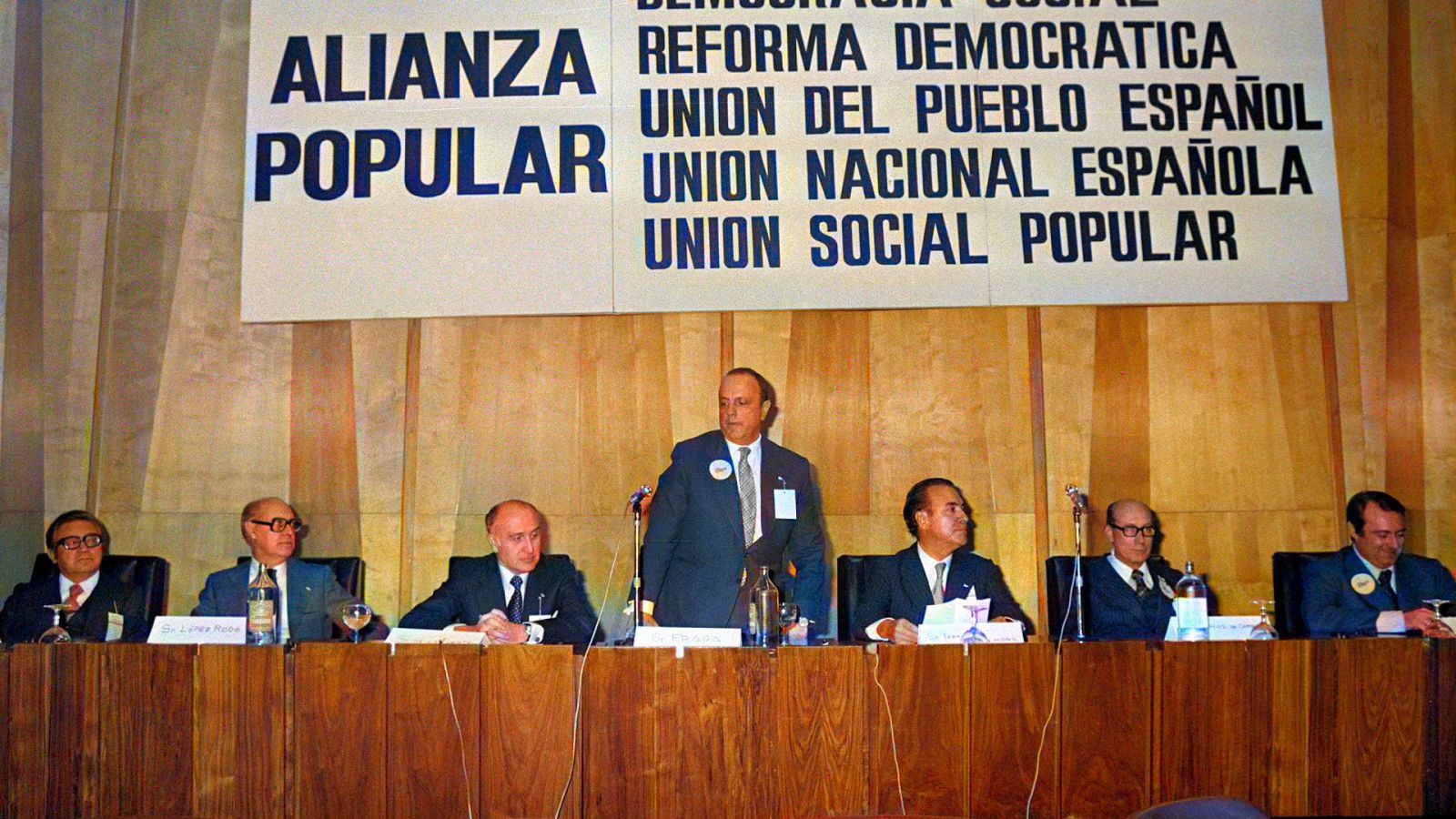 En la mesa presidencial, un hombre con traje oscuro y corbata habla por un micrófono, rodeado de otros hombres. Detrás, un cartel anuncia un evento político con varias consignas.