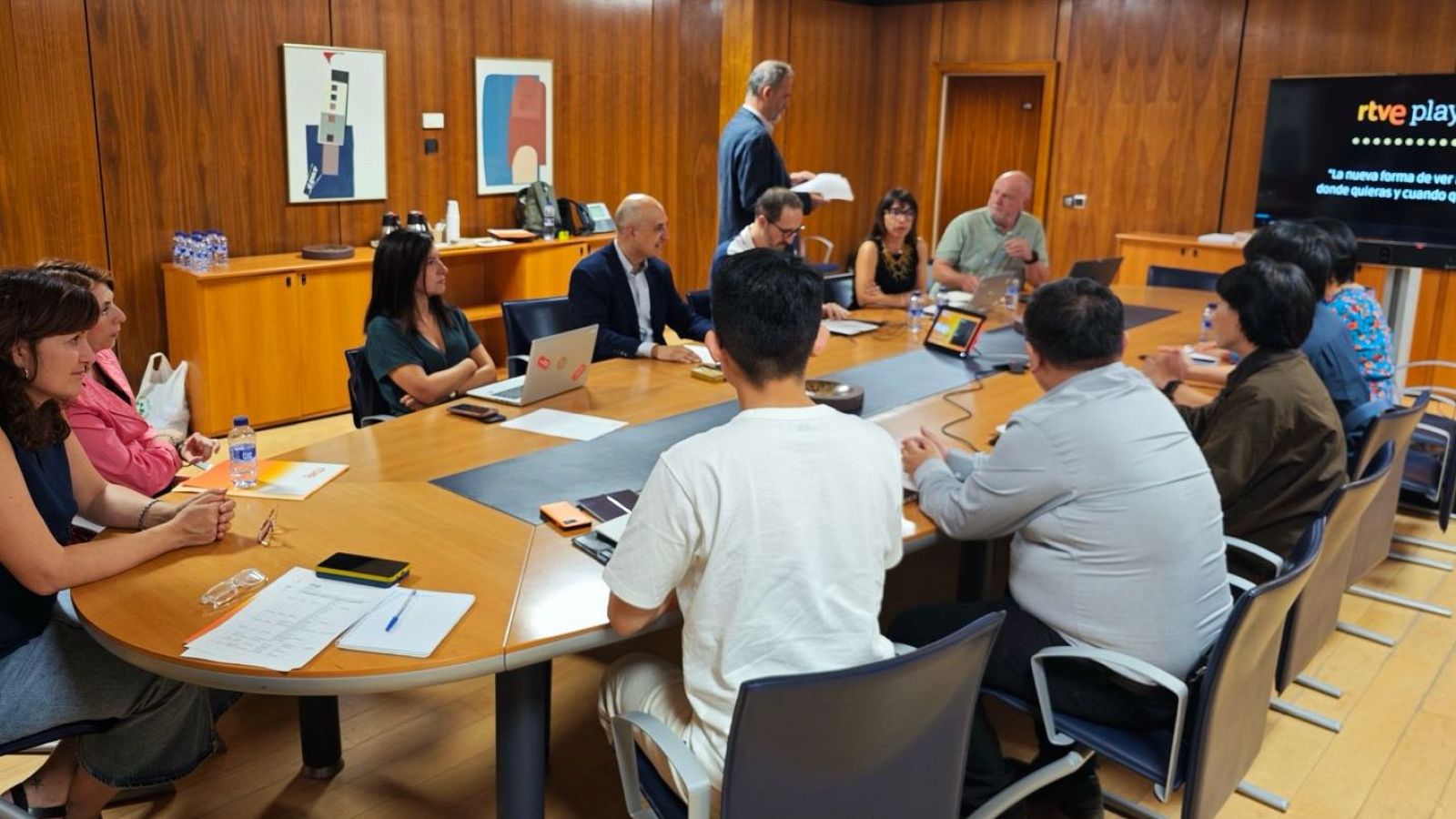 En una sala de reuniones, un grupo de personas se encuentra alrededor de una mesa ovalada. Algunos participantes toman notas y usan dispositivos electrónicos, mientras que una pantalla muestra un logo y un mensaje.