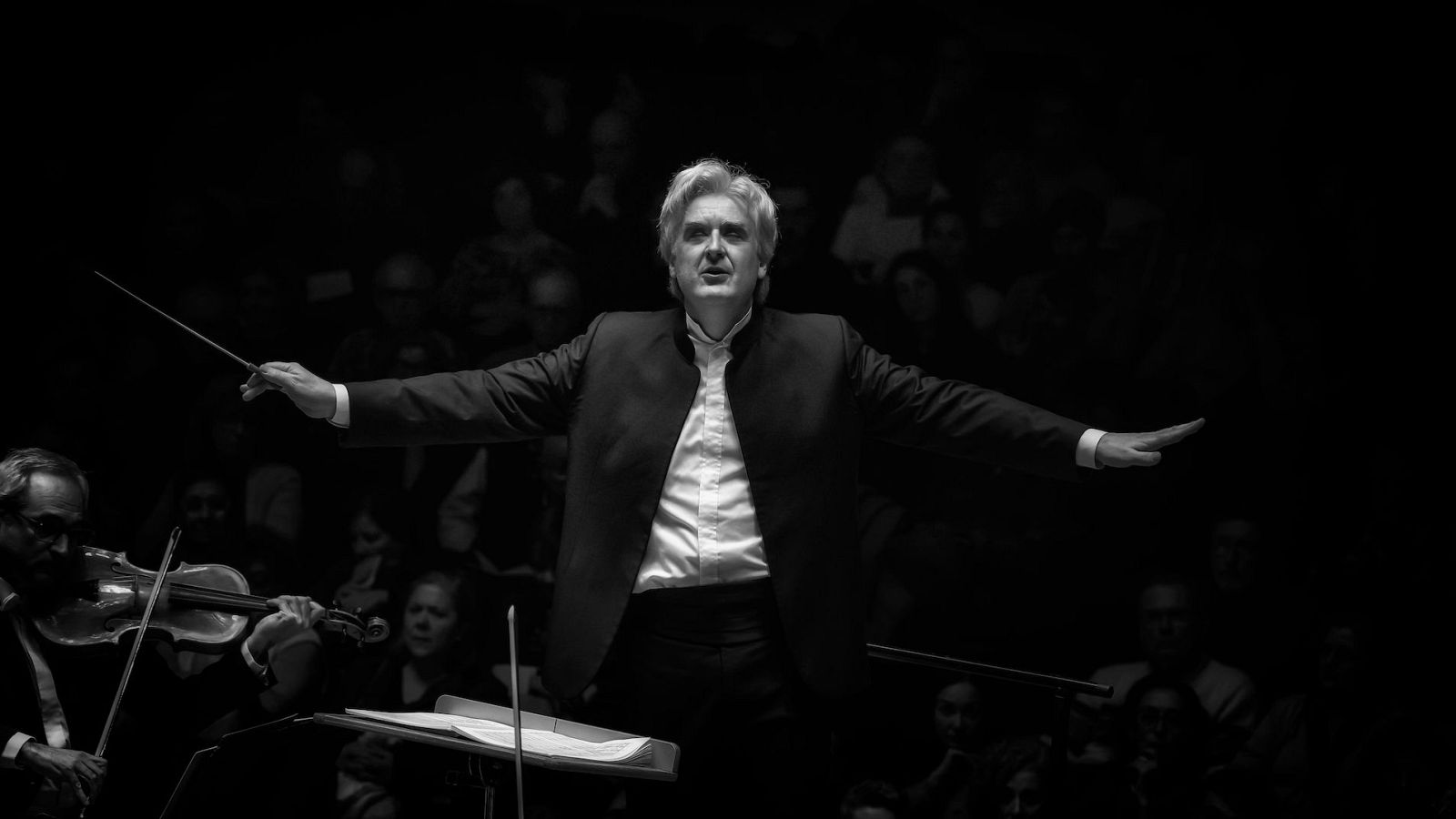 Thomas Dausgaard dirige a la orquesta, con una batuta en la mano, mientras un violinista toca en primer plano y el público observa el concierto.