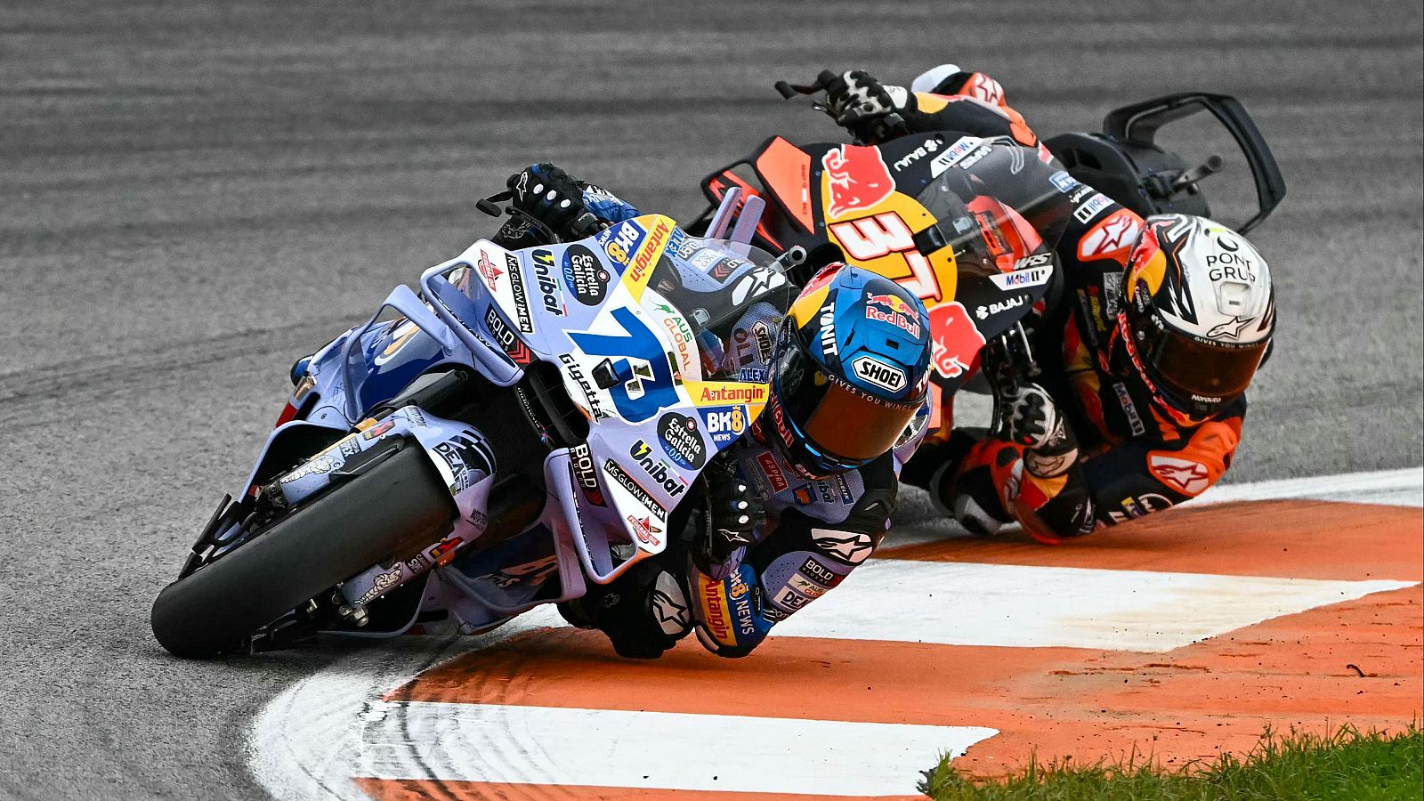 Dos pilotos de MotoGP compiten en una curva del circuito. Uno lleva una moto azul y blanca con el número 73, mientras que el otro pilota una moto naranja, negra y blanca.