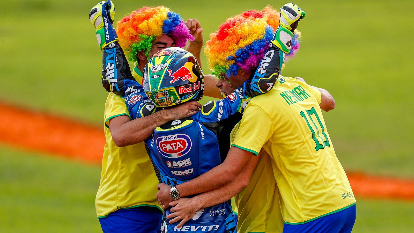 Diogo Moreira, con su casco y traje de carreras, es abrazado por dos personas con pelucas y camisetas amarillas con el nombre "Neymar" y el número "10", en una celebración con fondo verde.