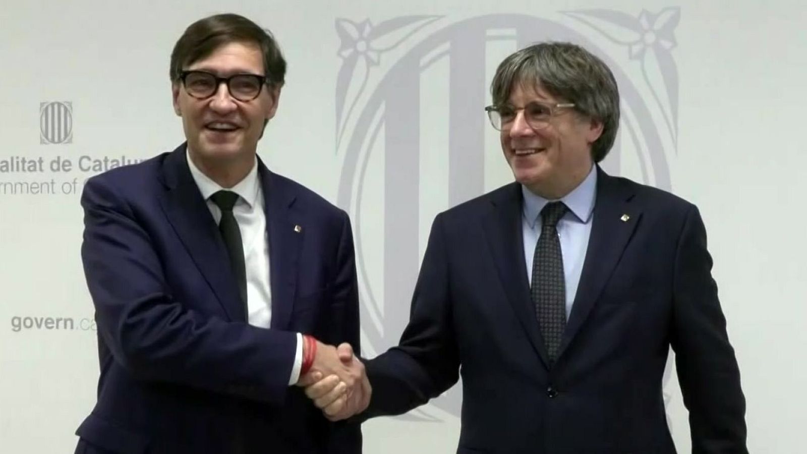 Puigdemont e Illa, en traje y corbata, se dan la mano en una reunión con el logo de la Generalitat de Catalunya de fondo.