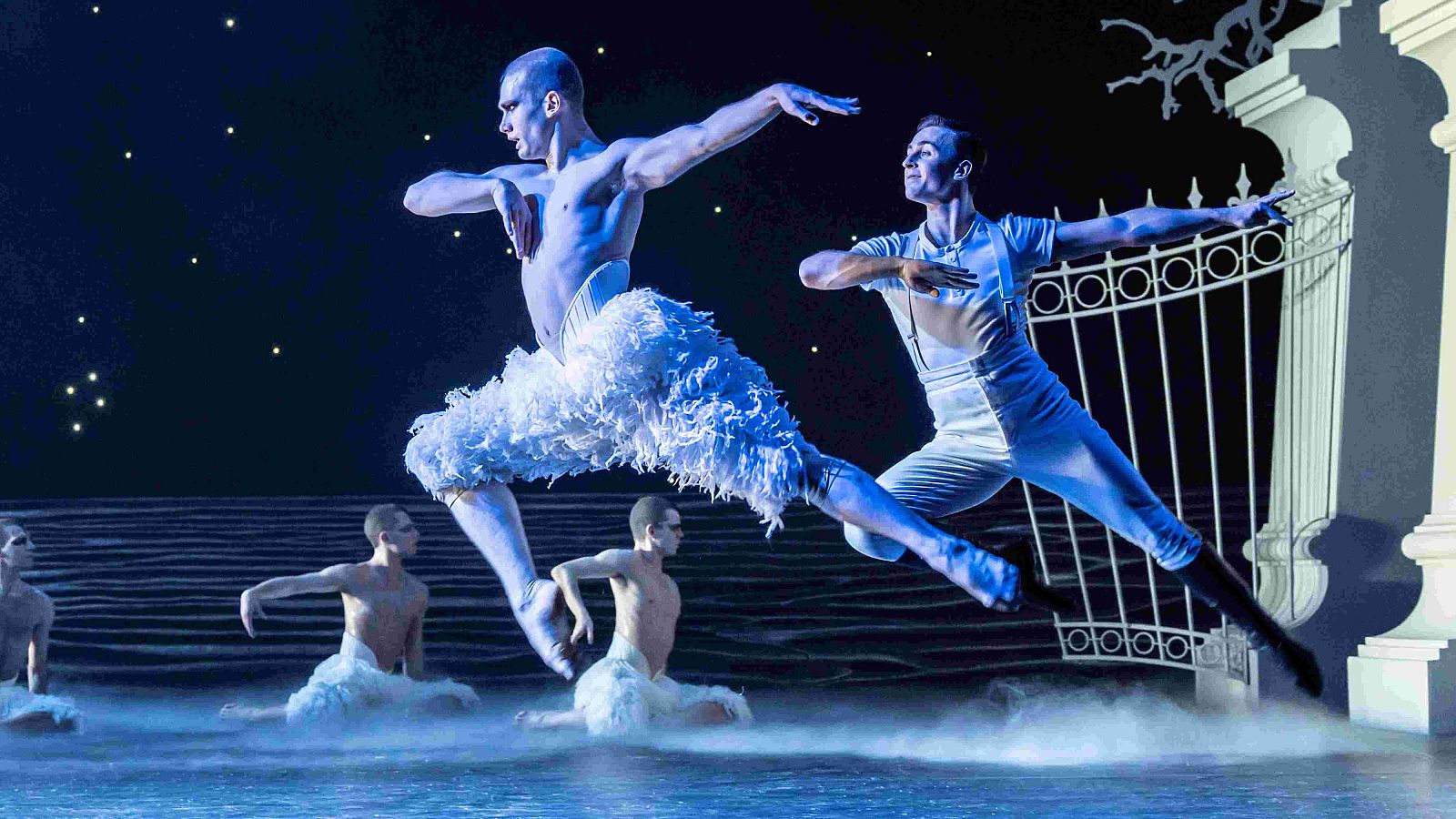 Dos bailarines masculinos ejecutan un salto de ballet en un escenario teatral, uno vestido de cisne con tutú blanco y el otro con traje blanco, mostrando gran agilidad y fuerza, con otros bailarines y una escenografía de fondo.