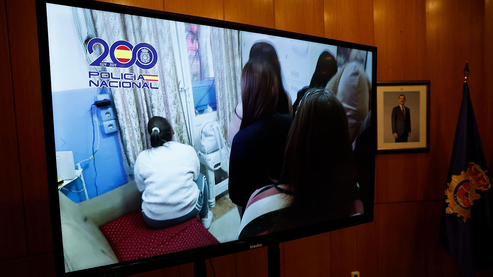 Una pantalla de televisión en una sala con paredes de madera muestra una imagen con el logo del bicentenario de la Policía Nacional y personas en una habitación, posiblemente víctimas de trata, con una persona sentada en una cama con colcha roja.