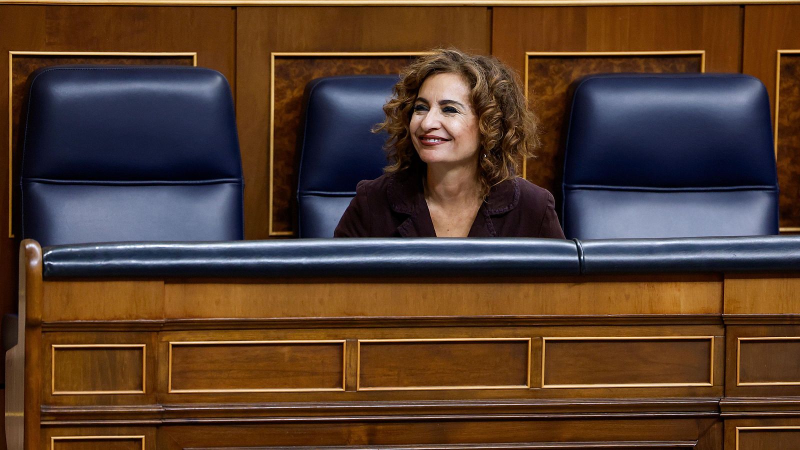 María Jesús Montero, Ministra de Hacienda, sonríe mientras está sentada en una sala de reuniones, posiblemente en el Congreso de los Diputados, frente a una mesa oscura y con sillas de cuero azul oscuro detrás.
