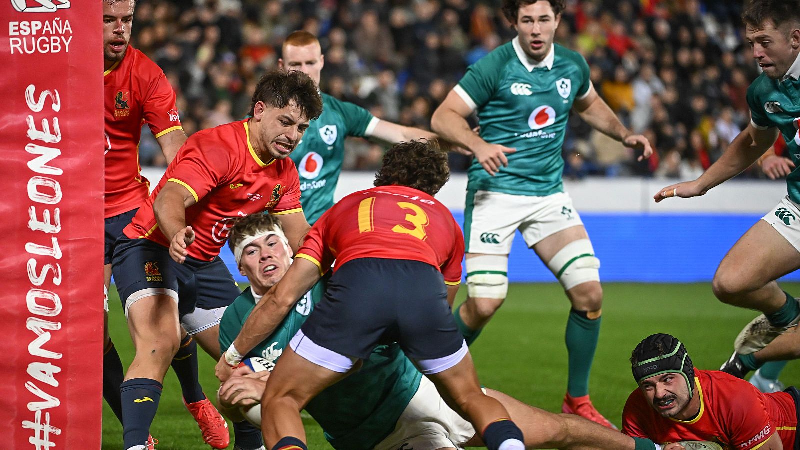 En un estadio lleno, un grupo de jugadores de rugby, vestidos con camisetas rojas y verdes, se enfrentan en una jugada intensa. La imagen muestra la fuerza y la estrategia del juego, con una pancarta de apoyo visible.